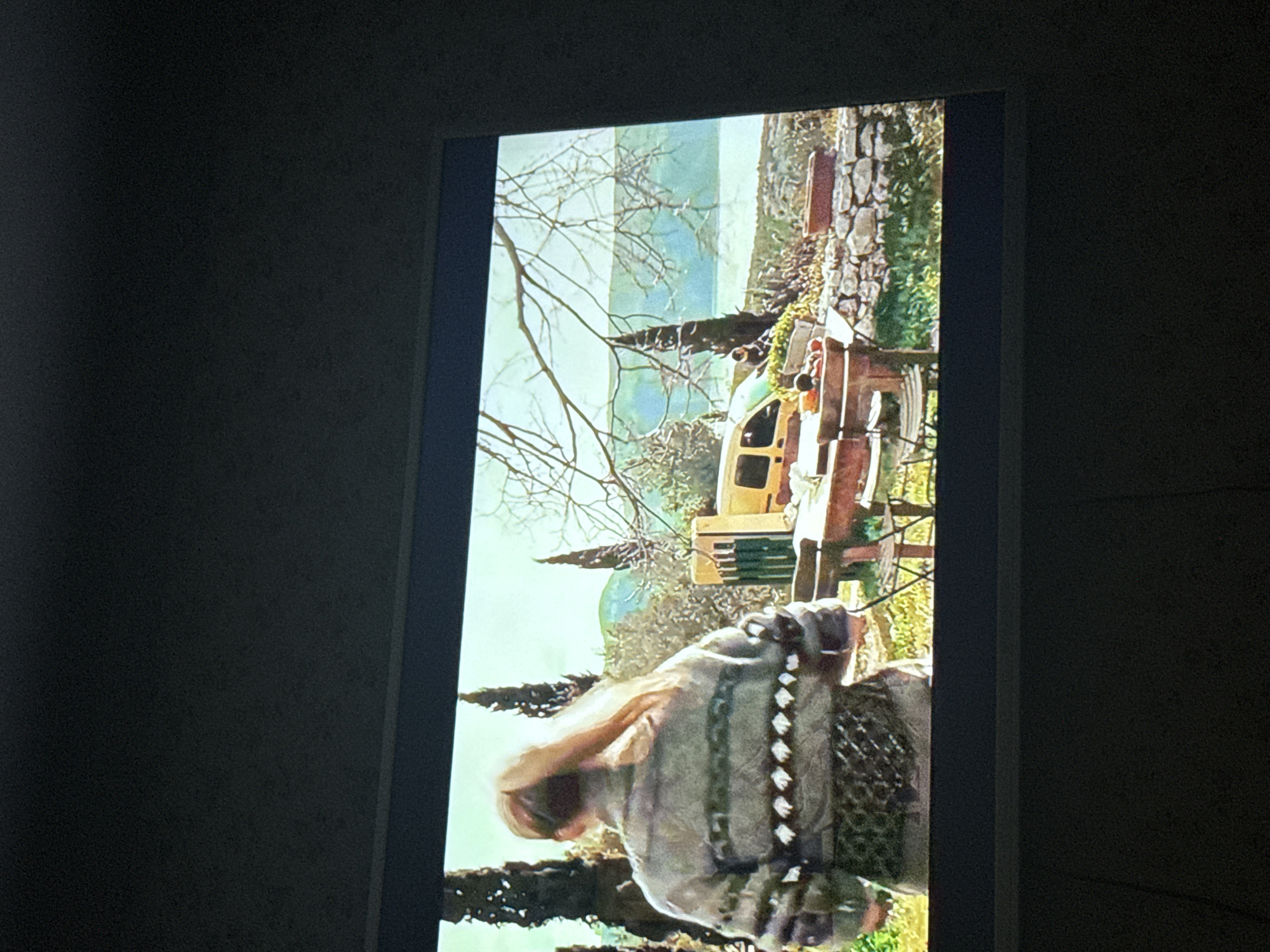 A photograph depicts a scene displayed on a screen, likely a television or monitor, set against a dark wall. The displayed scene appears to be an outdoor setting with a river or body of water visible in the distance. The foreground of the screen shows what seems to be a person wearing a textured garment, possibly a poncho or shawl, and there are also elements suggestive of a garden or natural area, including a table, chairs, and potentially a yellow vehicle. The scene includes what appears to be a stone wall on a grassy area with trees. The image provides no location information.