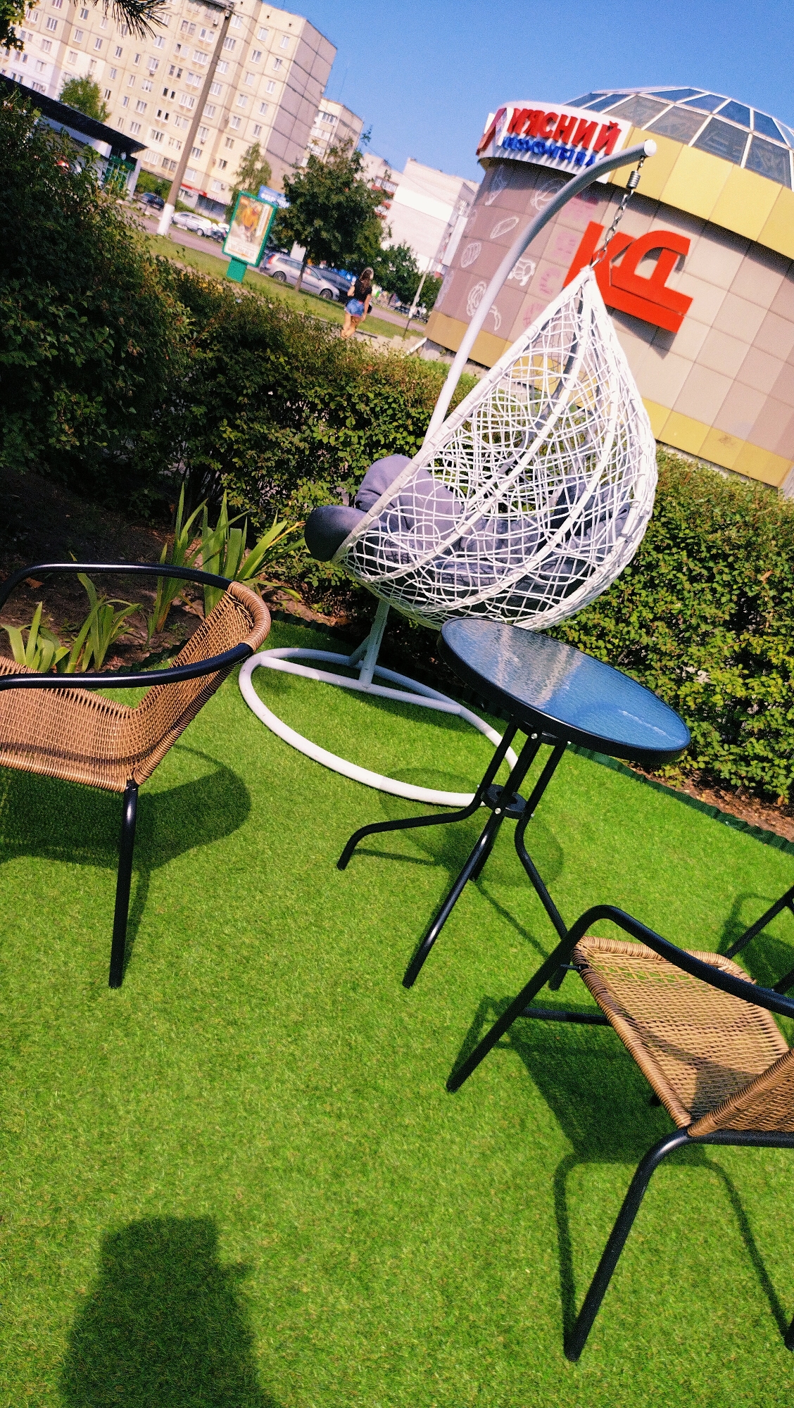 The image depicts an outdoor seating area with artificial turf, located in front of a shopping center in Horishni Plavni, Ukraine. The scene includes a hanging egg chair with a gray cushion, a small round glass-top table with a black metal frame, and two wicker chairs with black metal frames. In the background, a woman in a black top and denim shorts is walking on a sidewalk in front of a large, multi-story apartment building. The shopping center in the background features a large red "KC" logo and a domed glass roof. The sun is shining brightly, casting shadows on the artificial grass.