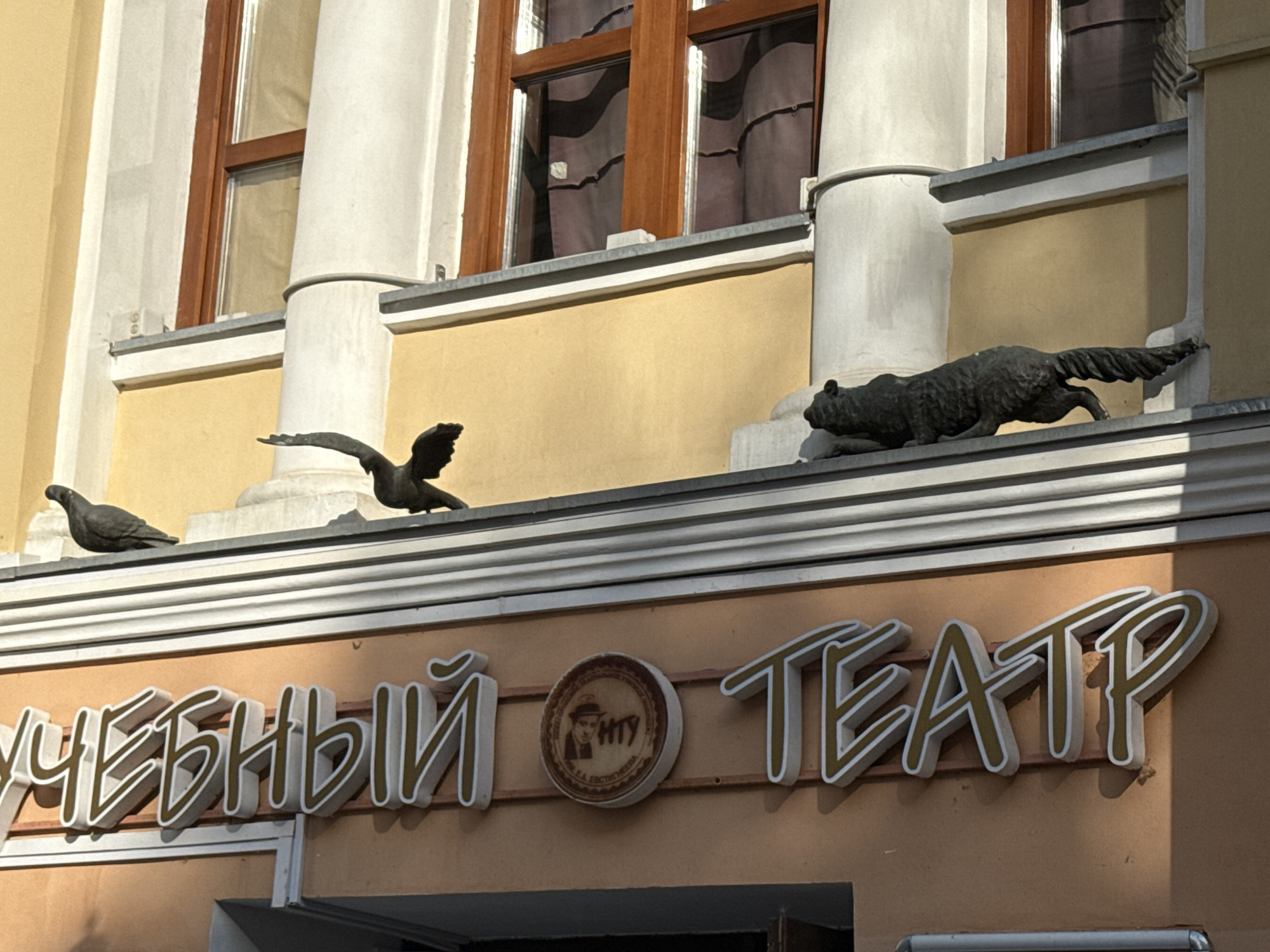 This is a medium-shot, outdoor, eye-level photograph of a building facade with a sign. The building appears to be historic, with architectural details such as columns and arched windows. The facade is painted a light yellow and beige.
On a ledge below the windows, there are two sculptures of birds. One is in flight, with its wings outstretched, and the other is perched. Further to the right, on a higher ledge, is a sculpture of a wolf or dog-like animal in a stalking pose.
Below these sculptures, a large, three-dimensional sign with white lettering and a gold outline reads, "УЧЕБНЫЙ ТЕАТР" (which translates to "Training Theater"). In the center of the sign, between the two words, is a circular emblem featuring a portrait of a man in a hat, with the Cyrillic letters "НТУ" visible. The emblem also has text around it, but it is too blurry to read.
The lighting suggests it is daytime, with shadows indicating the sun is at an angle. The scene is likely in a city given the building's architecture and the presence of the theater sign. The overall impression is one of a cultural institution with artistic embellishments.