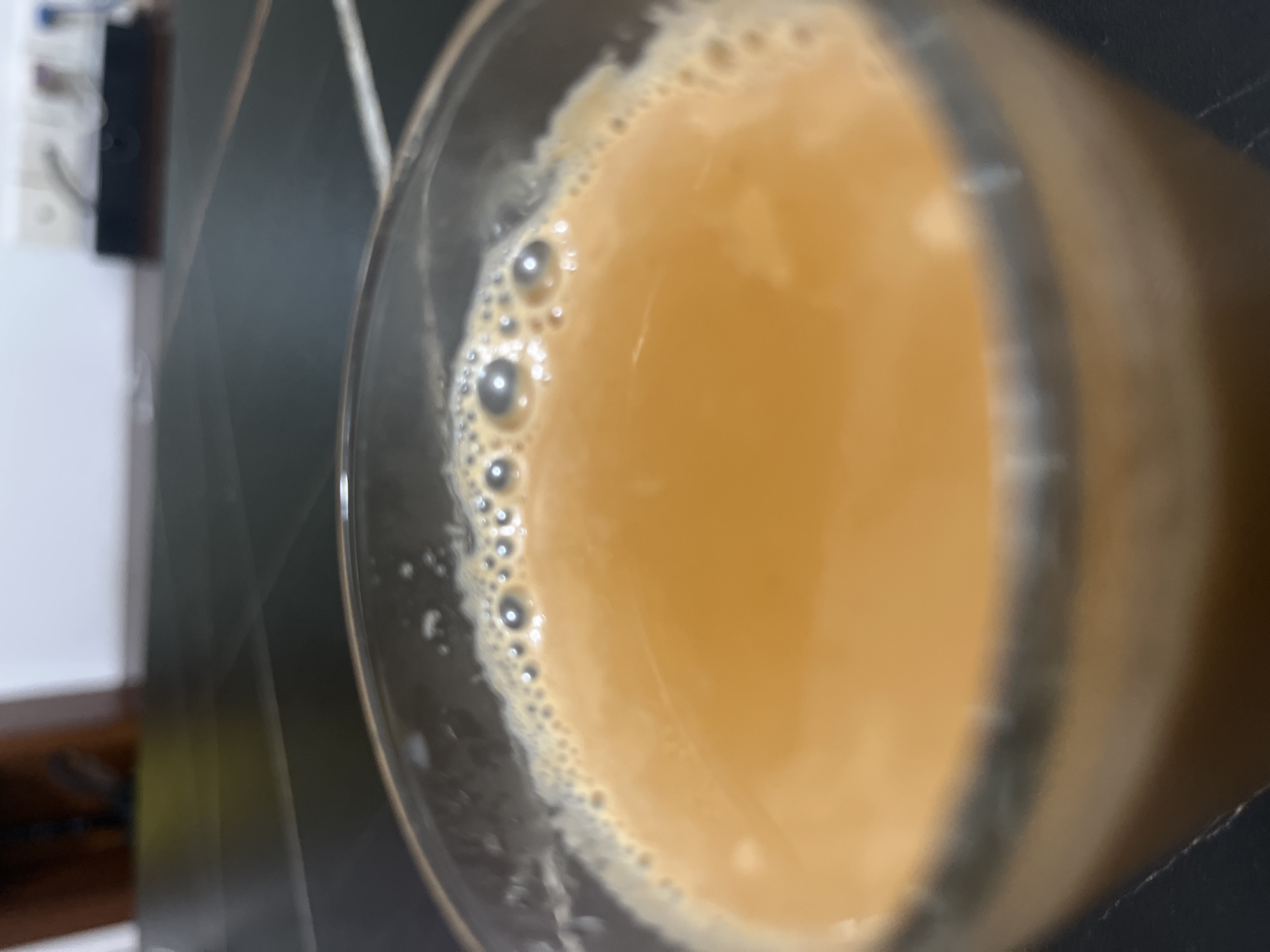 The scene is located in Jiulongpo District, China. The primary subject is a clear glass, viewed from above and slightly angled, containing a light amber or milky brown liquid. The surface of the liquid is topped with a layer of small, white bubbles, varying in size, concentrated around the rim and spread across the center. The glass appears approximately two-thirds full and rests on a dark, possibly black or dark grey, tiled or paneled surface that recedes into a blurred background. The background consists of a light-colored, likely white, wall situated behind the dark surface. On the right side of the background, blurred objects resembling electrical outlets or switches with connected cords are visible on the wall, positioned above a dark, rectangular object on the surface. On the left side of the background, a blurred dark brown object, possibly an item of furniture, is partially visible. No individuals or discernible actions are depicted in the image. The lighting suggests an indoor setting.
