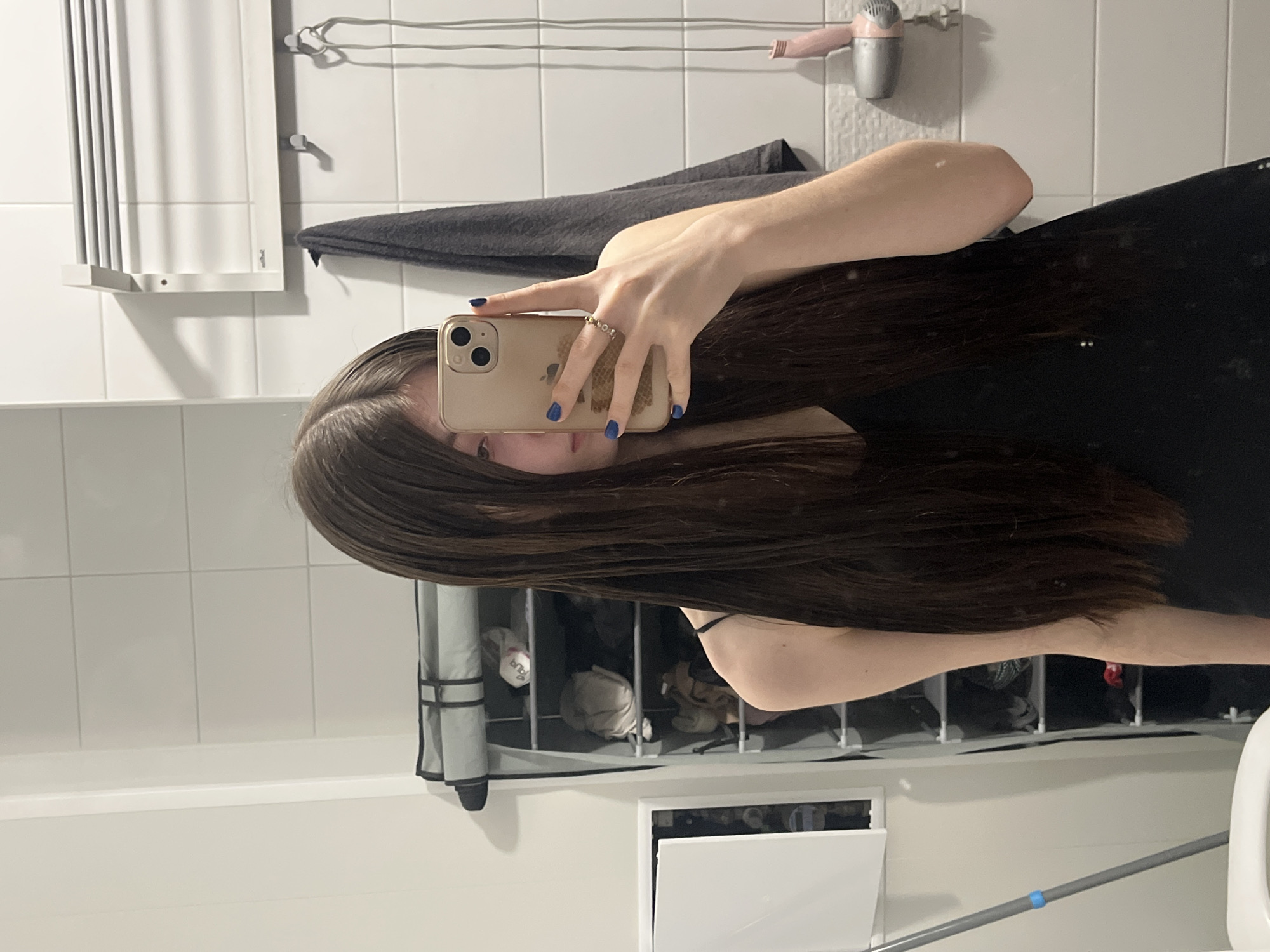 A person is holding a phone, presumably taking a selfie, in a bathroom. The background shows white tiled walls, a towel rack with a gray towel, a hairdryer, a storage unit with items inside, and a toilet. The location cannot be determined from the image.