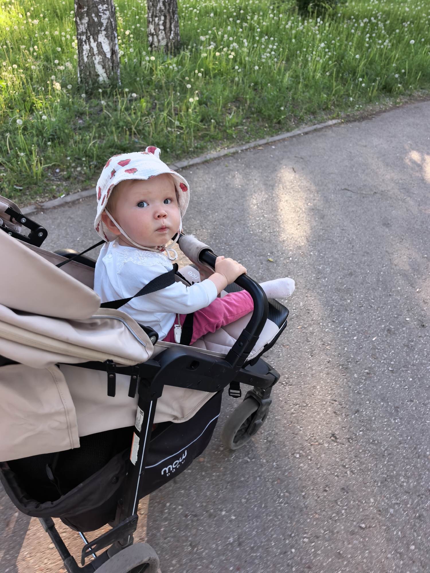 A fair-skinned baby sits in a black and beige stroller with pink pants, a white long-sleeved shirt, and a white hat patterned with strawberries. The baby is wearing a bib and looking forward. The stroller is on a paved path, likely a sidewalk, next to a grassy area with dandelions and trees in the background. The photo appears to be taken outdoors during the daytime with ample sunlight. The location may be Sterlitamak, Russia.