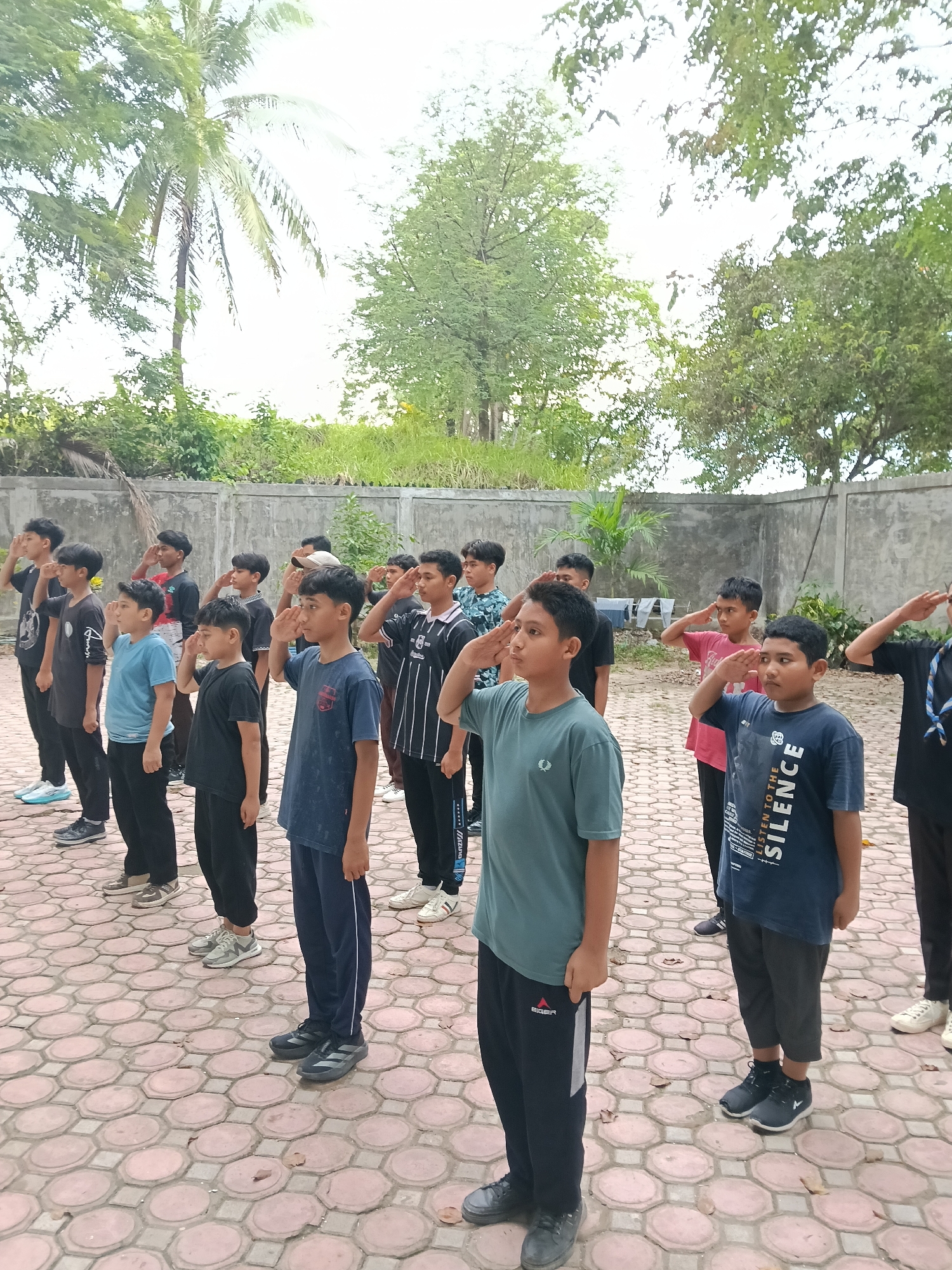 A group of approximately 15 young boys are standing in rows on a paved, hexagonal-tile surface, raising their right hands in a salute. The boys are wearing various casual shirts and pants; some are in tracksuits, and one has a blue neckerchief. In the background is a light gray concrete wall, behind which are trees and foliage, including a palm tree on the left side. The image was likely taken outdoors in daylight. The location appears to be the city of Lhokseumawe, Indonesia.