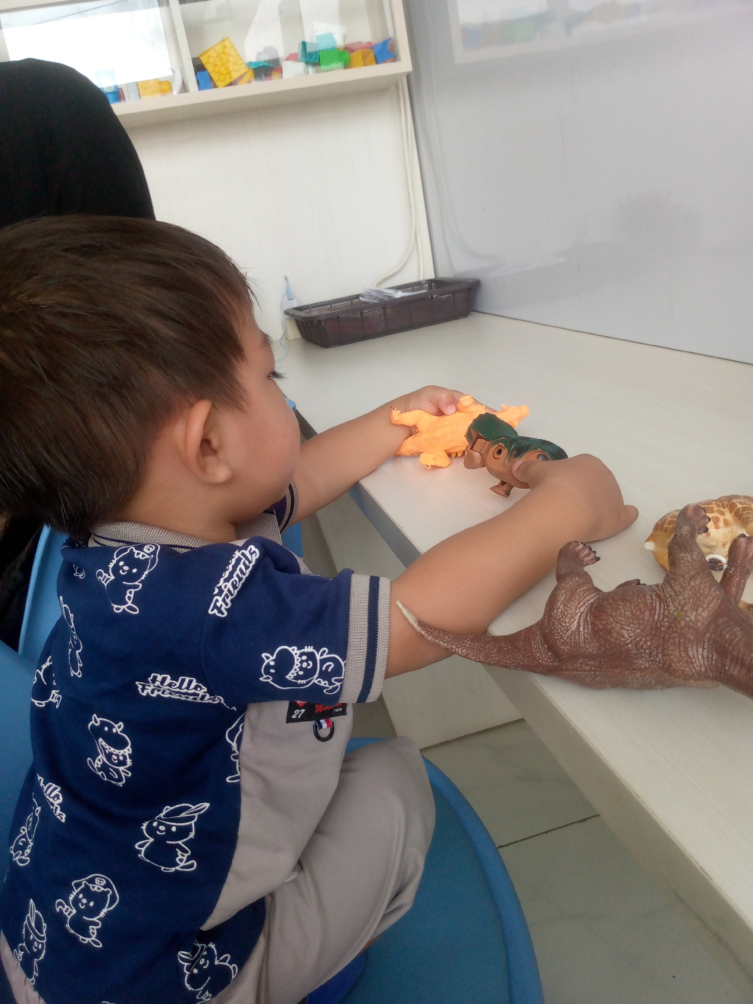A toddler is seated at a table, playing with several toy animals, including a dinosaur and what appears to be a crocodile and a giraffe. The toddler is wearing a navy blue shirt with white cartoon animal graphics and light grey pants. The table is white, and the background shows a shelf with colorful toys and a white board. The scene is lit by overhead lighting. The location is Kertajaya, Indonesia.