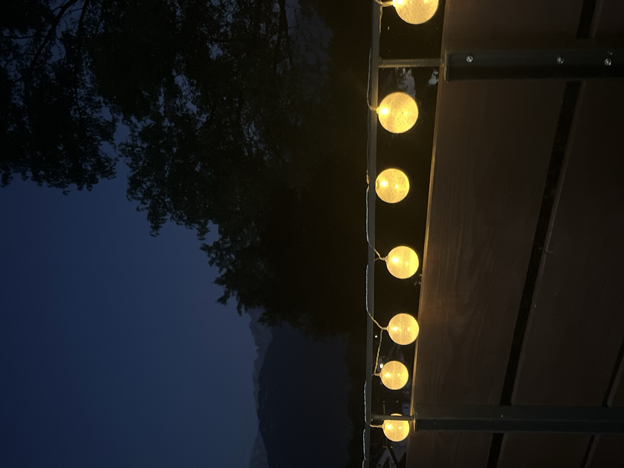 The image depicts a string of spherical, illuminated lights against a dark sky with faint mountain silhouettes in the background. The lights are yellow and appear to be suspended above a dark wooden railing, supported by vertical metal posts. The scene suggests an outdoor setting at dusk or night, possibly on a balcony or patio. The overall atmosphere is serene and ambient.