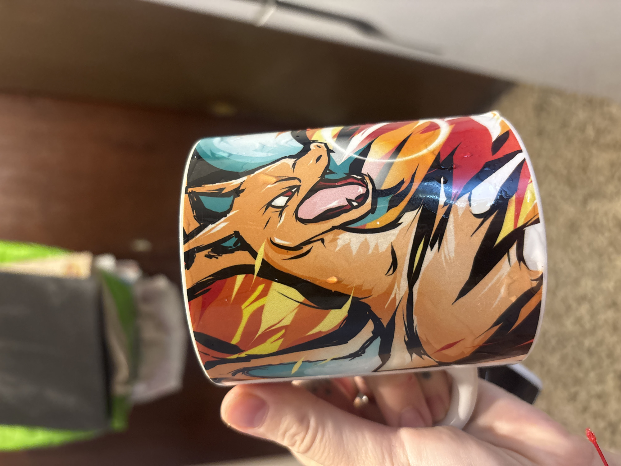 A person is holding a cylindrical mug with a glossy finish, featuring artwork of the Pokémon character Charizard against a backdrop of flames and teal accents. The mug is held by the handle with the fingertips visible. The background includes a blurred, dark wooden surface on the left and a light brown, textured surface on the right. The scene is indoors, lit by natural light. The image does not appear to be taken in Buenos Aires, Argentina.