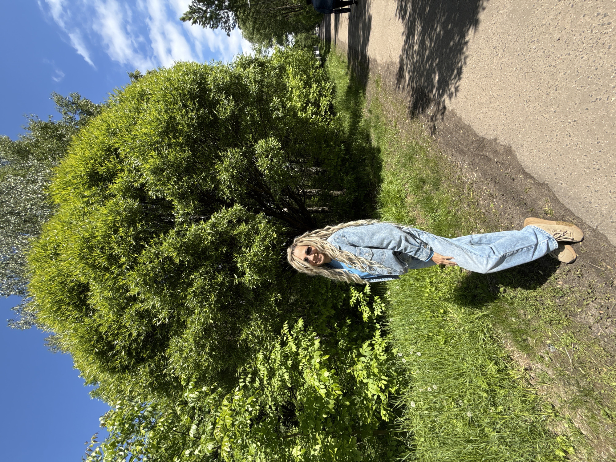 A young woman stands on a grassy patch beside a paved path in Заволжье, Russia. She is wearing a denim jacket and jeans, and beige shoes. The woman has blonde hair in long braids. Behind her is a large, round green bush, partially obscuring her. Above the bush is a clear blue sky with scattered clouds. The path to the right of the woman is partly shaded by trees.