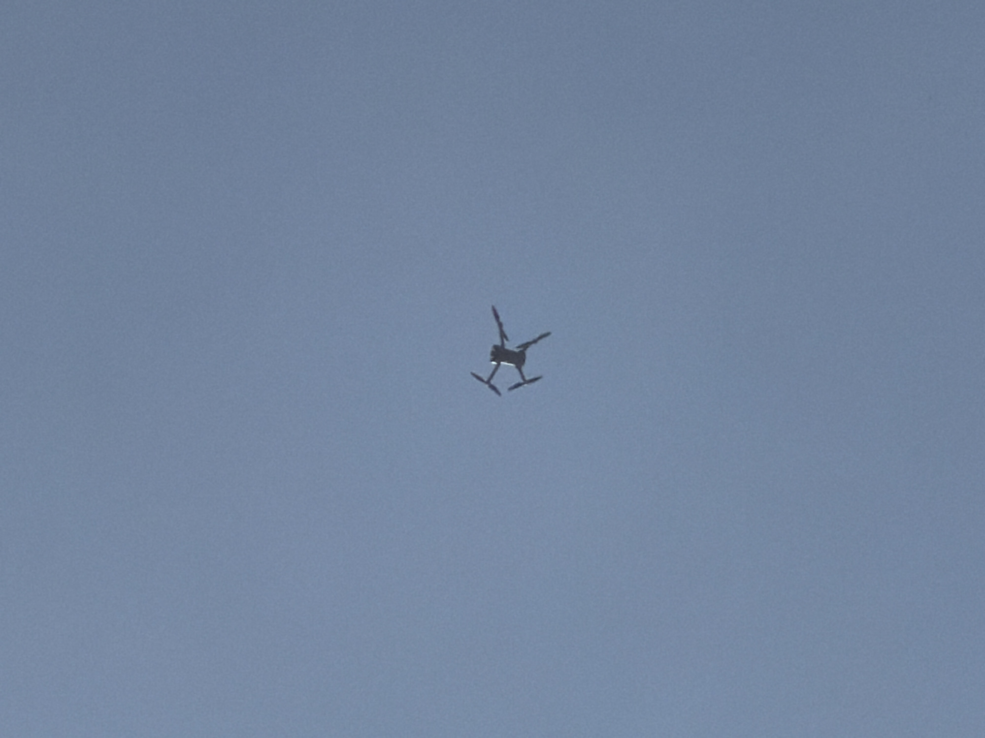 A quadcopter drone is depicted in flight against a clear, uninterrupted pale blue sky. The drone, appearing dark due to backlighting, is positioned centrally within the frame, showcasing its four outstretched propeller arms and central body from a perspective beneath it. No specific markings, model details, or discernible features other than its basic silhouette are visible on the aircraft. The sky serves as a uniform background, entirely devoid of clouds, land features, or any other environmental elements. The precise geographical location, including any specific area within the city of Bogdanitsa, Bulgaria, cannot be determined from the visual information presented in this image.