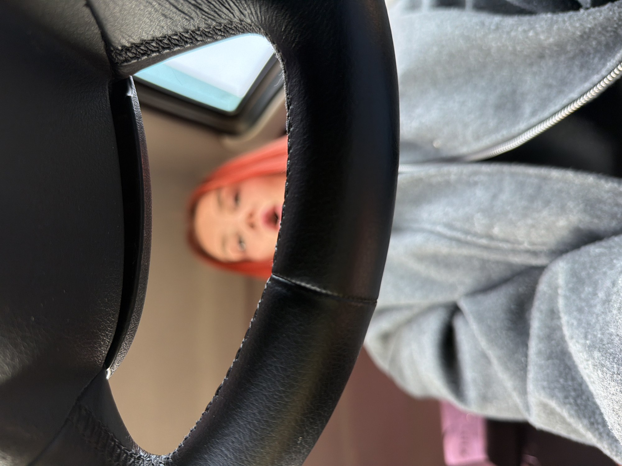 A close-up, low-angle shot through the car steering wheel. A person with red hair and wearing a gray zippered sweatshirt, face obscured, looks into the vehicle. A sunroof is visible above the steering wheel. The location is undetermined.