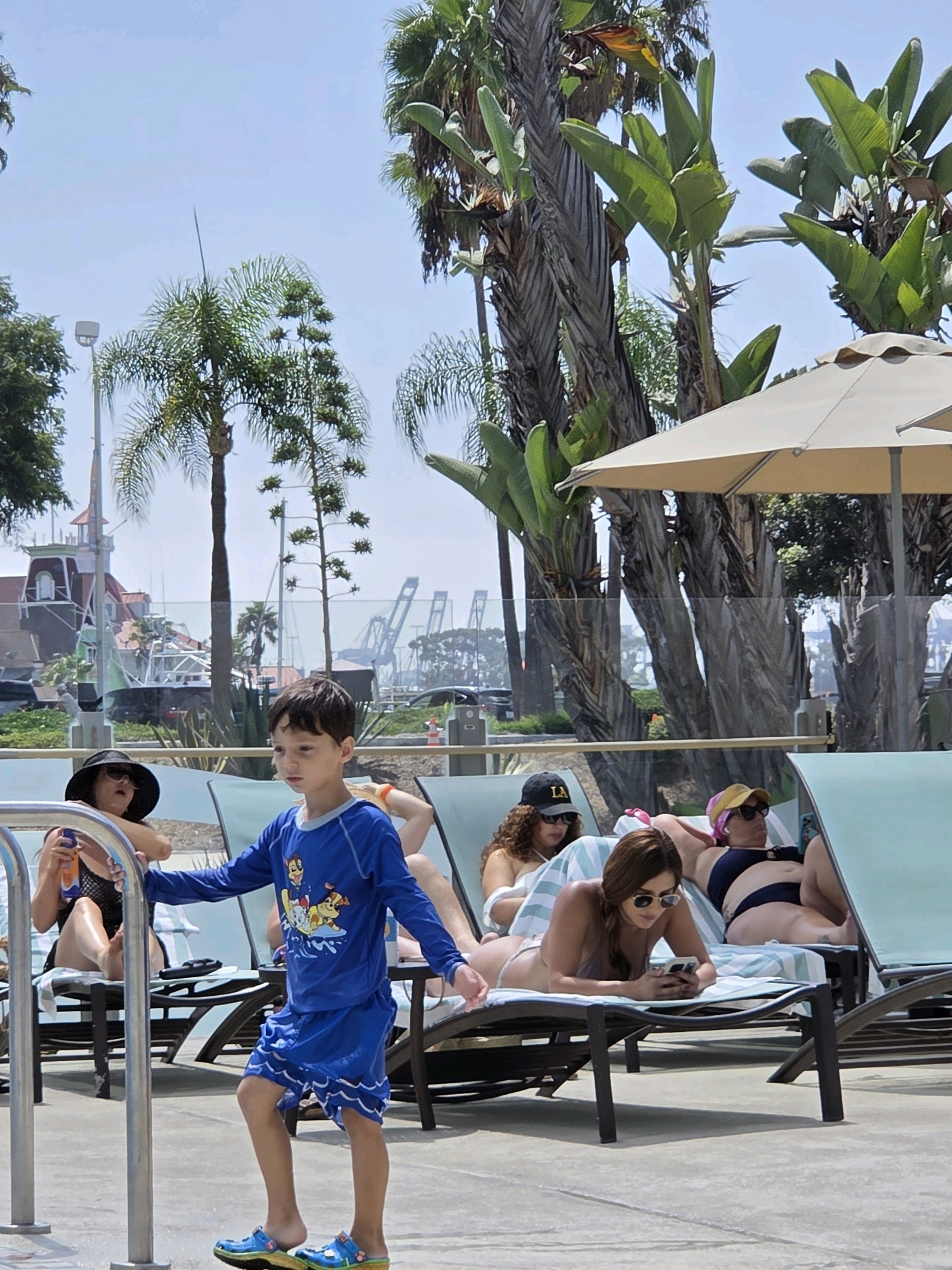 A boy in a blue rash guard and shorts with a Paw Patrol graphic is standing near a pool. He is wearing blue clogs. Behind him, several people are relaxing on lounge chairs by the pool. A woman in a black hat and swimsuit is holding a sunscreen bottle. Another woman in a yellow hat and sunglasses is lying on a lounge chair. A third woman in sunglasses is also on a lounge chair, looking at her phone. Palm trees and other tropical foliage are visible in the background, along with a hint of buildings and cranes, suggesting a waterfront or resort location. The sky is clear and blue, indicating a sunny day. The overall scene depicts a leisurely day by the pool on a warm day.