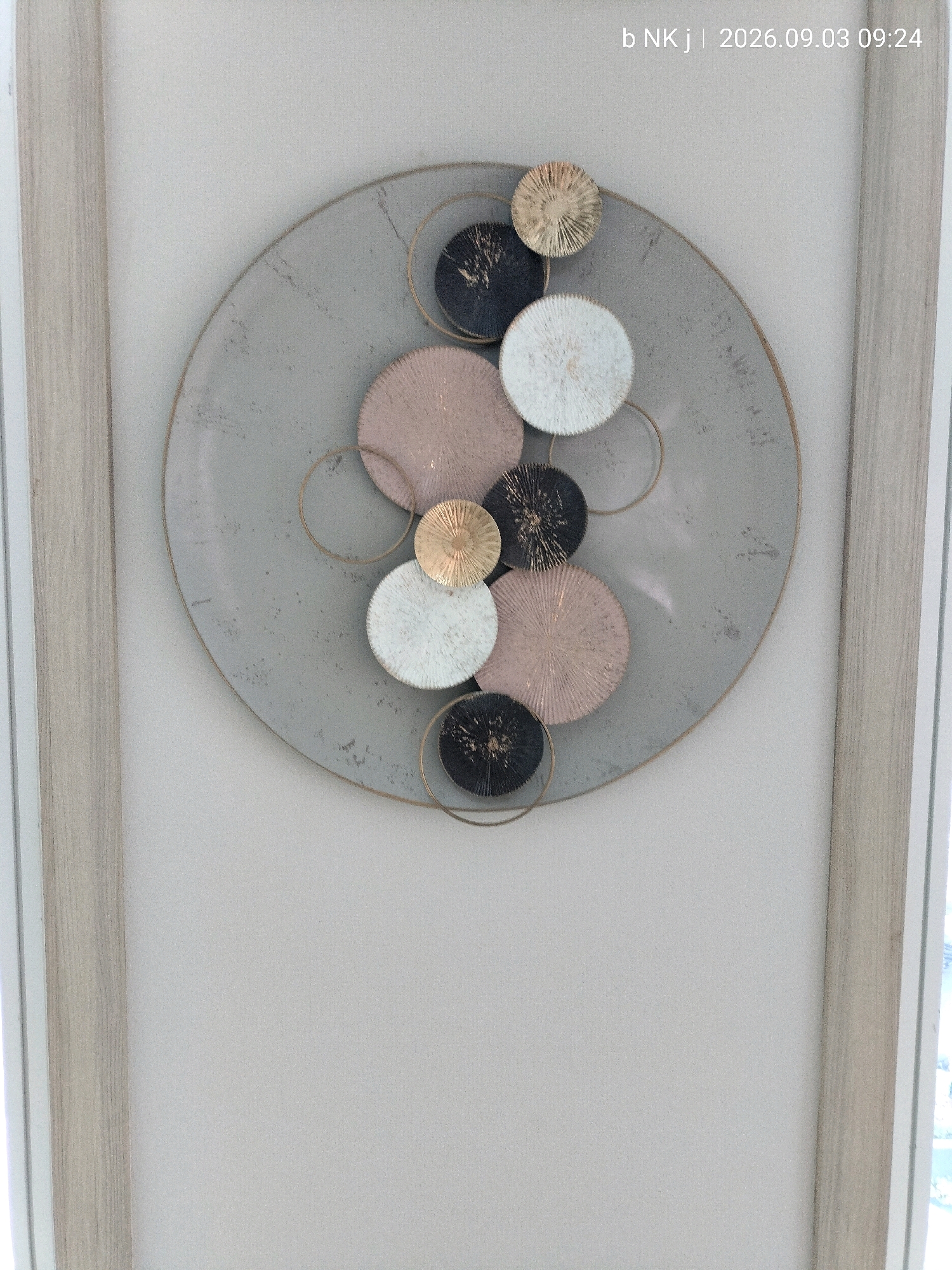 A circular, textured metallic wall art featuring multiple smaller, circular discs in varying colors and textures is centered on a light gray background. The discs are arranged in a radial pattern, with some overlapping. The colors of the discs include white, black with gold flecks, pinkish-gold, and gold. Several thin gold rings are incorporated into the design. The wall art is framed by vertical wooden elements on the left and right. A timestamp "b NK j | 2026.09.03 09:24" is visible in the upper right corner. The environment appears to be an indoor setting.