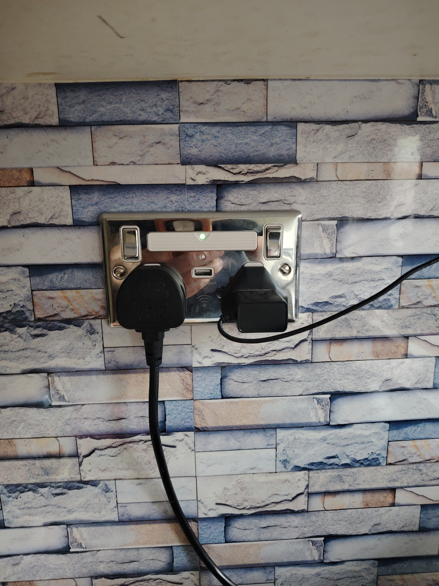 This image displays a chrome-finish double electrical wall socket with integrated USB charging ports, located in Huntingdonshire, United Kingdom. The metallic faceplate features two standard UK 3-pin outlets, each equipped with a polished toggle switch. A central white rectangular module, possibly a smart control unit, exhibits an illuminated green LED indicator. Below this module, a USB-A port and a USB-C port are present, with accompanying text indicating "2400mA" and a Wi-Fi symbol. A black UK 3-pin plug is inserted into the left outlet, with its black power cable extending downwards. A smaller black power adapter is plugged into the right outlet, with its thinner black cable extending to the right. The polished surface of the socket reflects a distorted image of a person holding a mobile device. The wall surrounding the socket is adorned with a wallpaper or tiling pattern designed to mimic irregular stacked stone bricks in varied shades of grey, blue, and beige. Above the patterned wall, a plain, light-colored surface is visible.