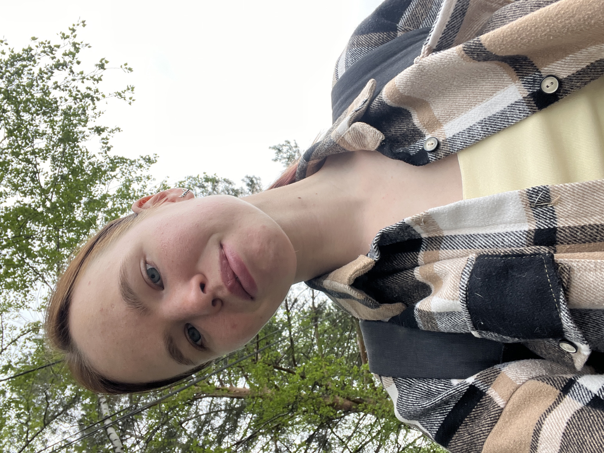 The photograph shows a person, likely a young woman, from a low-angle perspective with the camera looking upwards. The person has fair skin, light-colored eyes, and light brown or reddish-brown hair. They are wearing a plaid flannel shirt, which is primarily beige, white, and black. Underneath the flannel, there appears to be a yellow garment.
The background includes trees with green foliage and a cloudy sky, suggesting an outdoor environment, possibly a park or wooded area. Given the location metadata, it is possible the photo was taken in or near Chaikovsky, Russia.