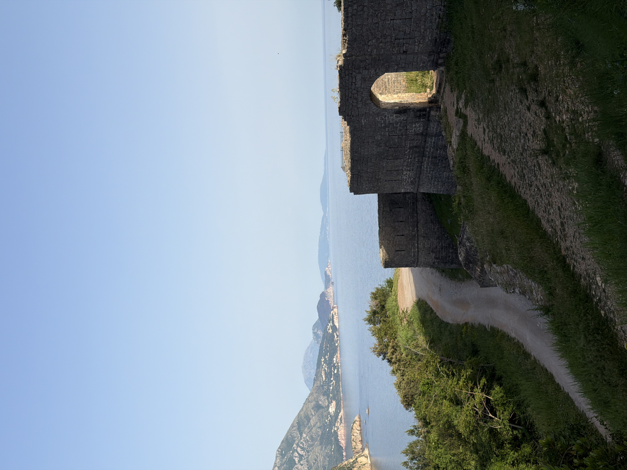 The image shows a view of the Adriatic Sea and coastline from a stone structure, likely part of the Citadel of Budva, located in Budva, Montenegro. The stone structure includes an arched opening and a pathway running alongside it. The sea is visible in the middle ground with distant landmasses. In the foreground, there is vegetation, including trees and grass. The sky is light blue.