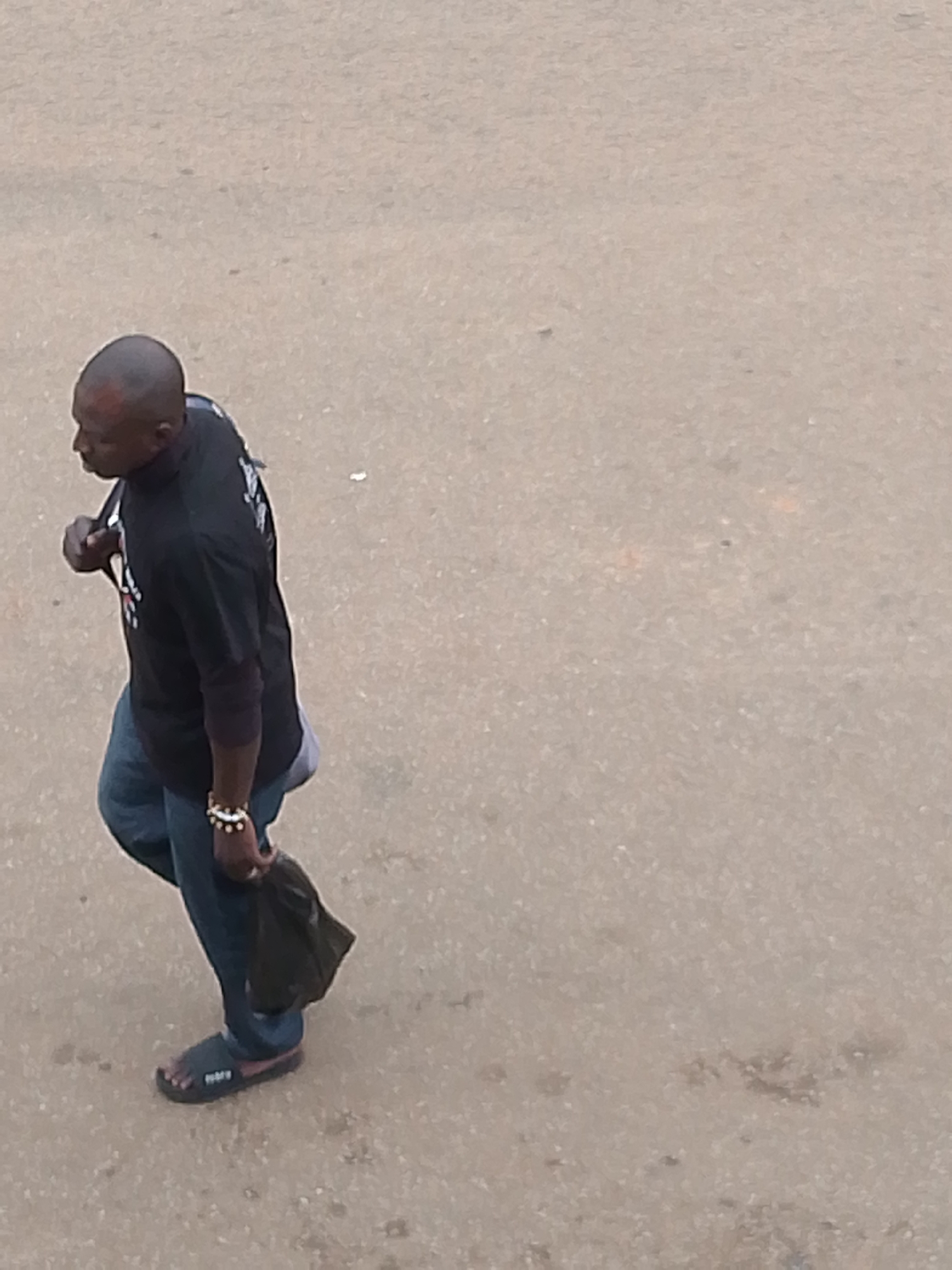 An overhead, medium-shot captures a Black man walking on a textured, pale gray surface that appears to be asphalt or concrete in Bukuru, Nigeria. The man is wearing a black t-shirt with white graphics on the front and dark blue jeans. He has a dark, short haircut and is wearing a bracelet on his left wrist. He is carrying a dark plastic bag in his left hand, which is dangling by his side. The man is wearing black flip-flops with a red sole. He is walking from the left of the frame towards the right. The background is an undifferentiated, textured surface with a few small specks of lighter and darker material visible.