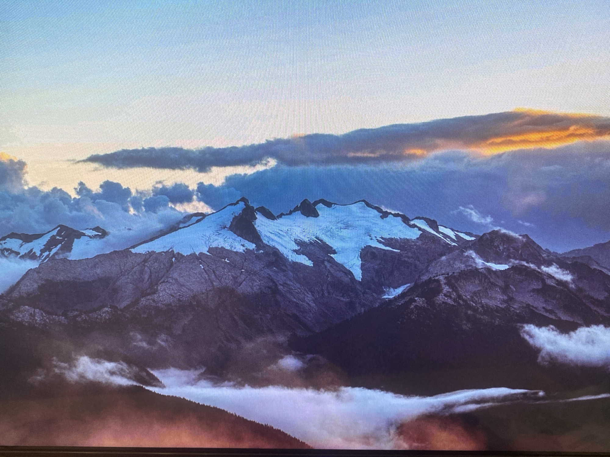 The image presents a majestic mountain landscape, likely captured from a digital screen due to a discernible pixel grid pattern. The scene features a vast mountain range with prominent, snow-capped peaks and glaciers, contrasting with the dark, rugged rock formations. The sky above transitions from light blue to deeper blues and purples, with a striking band of clouds in the upper right illuminated with warm golden and orange light, indicative of either a sunrise or sunset.
Clouds and fog are an integral part of the scenery, present at various altitudes. Some dense clouds drift across the upper sky, while others are nestled among the mountain peaks and cling to their slopes, partially veiling the intricate details of the rock faces. A thick layer of low-lying mist or fog fills the valleys and lower elevations, creating an ethereal atmosphere. Through this mist, the dark silhouettes of forested hills or slopes are faintly visible in the foreground, with some areas glowing with a reddish-brown hue from the indirect light.
There are no people, animals, or artificial structures visible, accentuating the wild and untouched nature of the environment. The overall impression is one of immense natural beauty, tranquility, and grandeur. While the location context mentions Saint Petersburg, Russia, the mountainous, glacial landscape shown is not characteristic of that region, which is generally flat. The image itself provides no specific geographical markers. No text is visible in the media file.