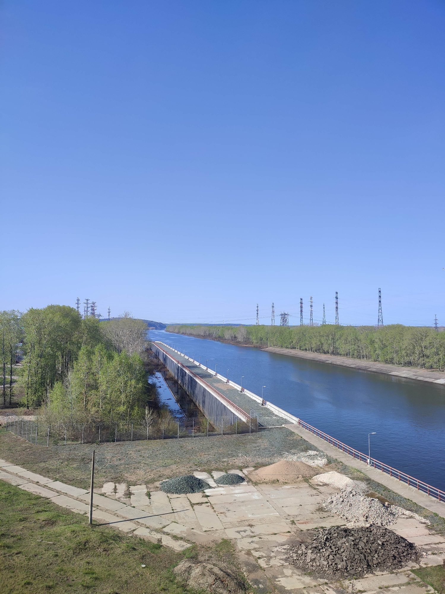 The image shows an industrial canal landscape under a clear blue sky. A canal, flanked by trees on either side, stretches from the foreground towards the horizon. A long bridge runs across the canal, appearing to connect different sections of the industrial area.
In the foreground, there is a concrete area with piles of gravel and debris, suggesting construction or maintenance activities. A weathered post stands near the edge of the concrete, close to a grassy area. The concrete is broken in several places.
Power lines are visible in the background, further indicating the industrial nature of the location.
The location is Unknown, Unknown. The overall scene suggests a quiet, industrial zone with a focus on water management or transportation.