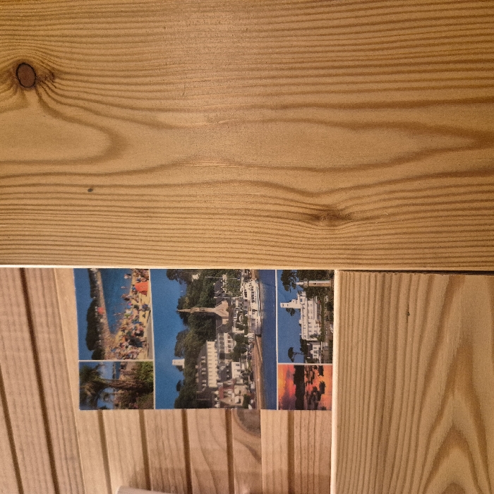 A postcard depicting various scenic views is partially visible against a backdrop of wooden planks. The postcard features images of a beach filled with people, a coastal town with white buildings, a large white building by the water, and a sunset. The wooden planks above and below the postcard have a light brown color with distinct grain patterns.