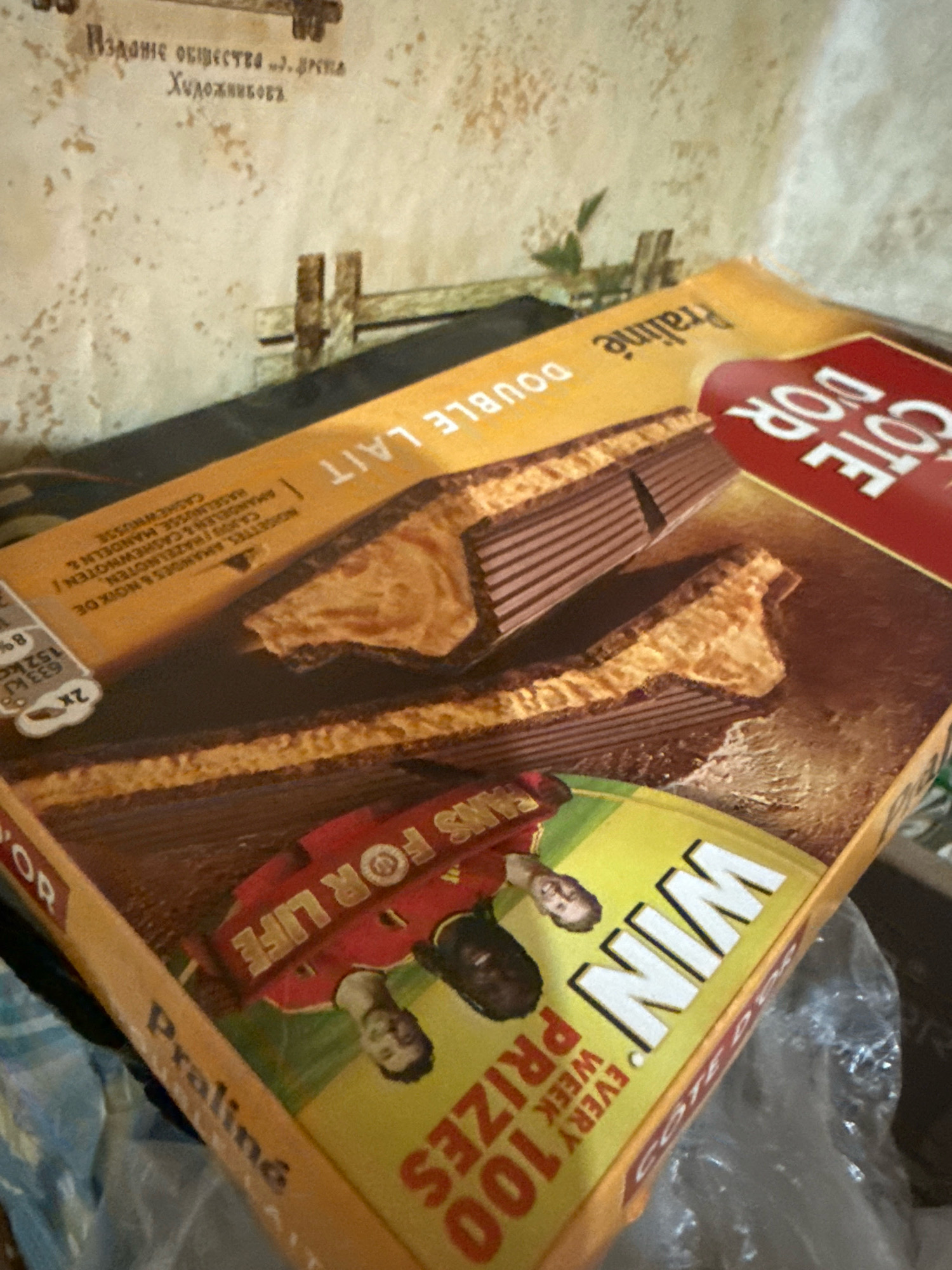 The image shows a close-up of a box of Cote d'Or Praliné Double Lait chocolate bars. The box is made of cardboard and is yellow and red. The chocolate bars are layered and appear to be filled with a praline cream. The box has text in French, including "Praliné Double Lait" and "Cote d'Or". There is also text and graphics related to a promotion, with "WIN 100 PRIZES EVERY WEEK" visible. The background is a textured wall, possibly a plaster wall, with some faint writing in what appears to be Cyrillic script. There are also some wooden elements in the background, possibly part of a fence or a shelf. The overall scene suggests the product is being displayed or stored. There is no information in the image that would allow for the identification of its location in Basarabeasca, Moldova.