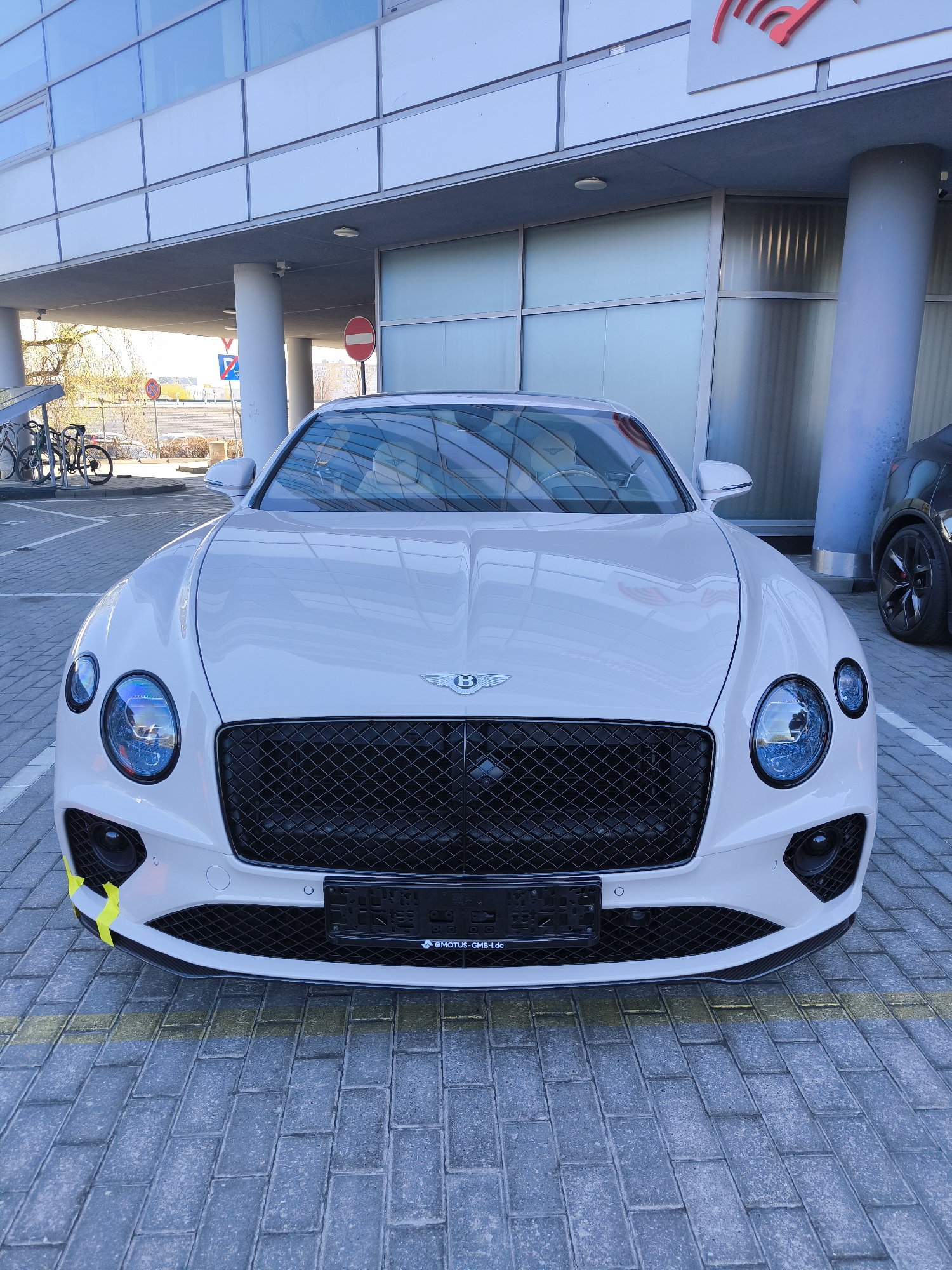 A pristine white Bentley car is prominently displayed in a head-on view, parked on a paved surface. The car features a large, black mesh grille with the iconic "B" emblem centered, flanked by large circular headlights with a distinct blueish tint and smaller auxiliary lights. A yellow accent or piece of tape is visible on the lower left side of the front bumper. The license plate holder at the bottom of the grille reads "EMOTUS-GMBH.de".
The car is situated in what appears to be a modern parking area or an entrance to a contemporary building. The ground is made of grey interlocking pavers, with a yellow line marking the parking space. In the background, a building with extensive glass windows, a light-colored facade, and a large concrete overhang supported by cylindrical pillars is visible. A red logo, possibly part of a business sign, is mounted on the building's exterior.
To the left of the car, under the building's cover, a bicycle rack holds several bicycles. Further back, road signs, including a red circular 'no entry' sign and a blue directional sign, are discernible. On the far right, part of a dark-colored car, specifically its front wheel and fender, is visible. The scene is brightly lit by natural daylight, indicating it is daytime. The overall setting suggests a commercial or office environment.