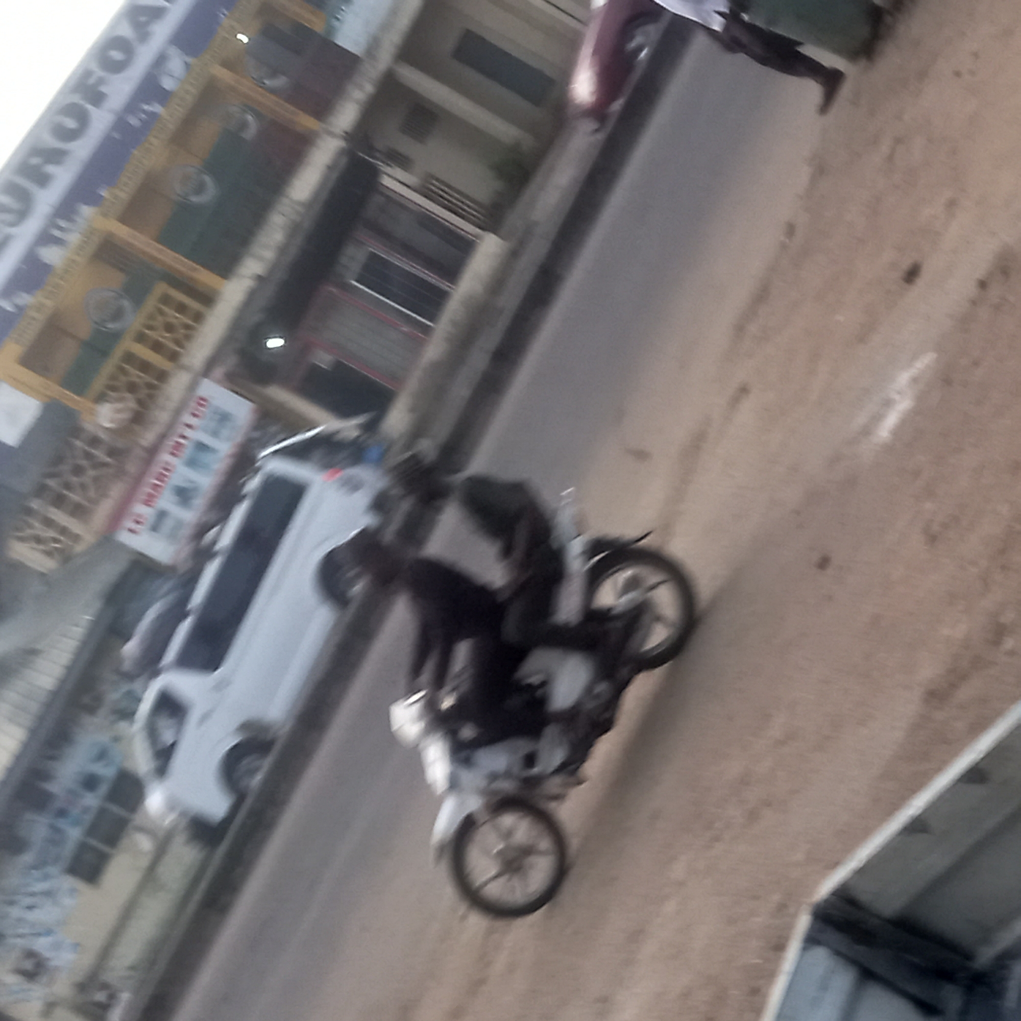 The image presents a tilted, somewhat blurry street scene in an urban or semi-urban environment, likely taken during the daytime. The primary subjects are a person riding a light-colored motorcycle, possibly with a passenger or cargo on the back, and a white van. The motorcycle appears to be in motion, as its wheels are blurred.
The activity taking place is typical street traffic. The motorcycle is positioned on what appears to be the road, while the white van is on the left side, either moving or parked near the curb. In the background, other people are faintly visible, and additional vehicles might be present.
The setting consists of buildings lining one side of the street. These structures vary in appearance, suggesting a mix of commercial and possibly residential uses. One building features distinct yellow-colored architectural elements, possibly wooden or metal grilles.
Notable details include:
- **Visible text:** On a large blue sign high up on one of the buildings, the word "EUROFOAM" is partially visible. On a white rectangular sign further down on another building, the text "LE MARCHÉ DU DÉTAIL" (French for "The Retail Market") can be partially read.
- **Time of day:** The lighting suggests it is daytime, without harsh shadows, indicating either an overcast sky or early morning/late afternoon.
- **Location cues:** The general style of the buildings and vehicles is consistent with scenes found in parts of Africa, aligning with the provided Sardauna, Nigeria context.
The image's blurriness and tilted perspective contribute to an impression of dynamism and everyday street activity.