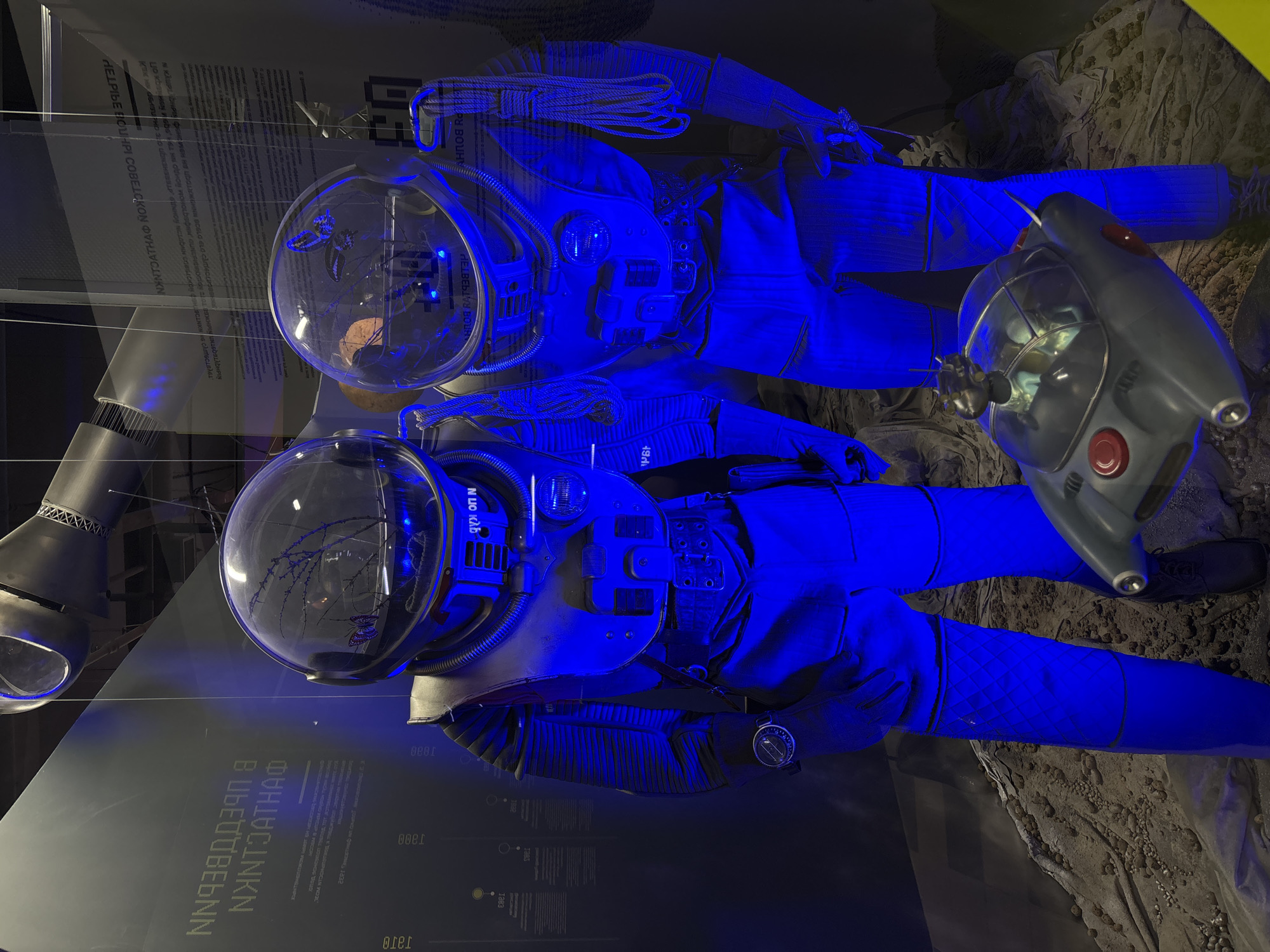 The image captures a display of two spacesuits, bathed in blue light, likely in a museum or exhibition. Each suit features a clear helmet with internal details and a complex control panel on the chest. The suits appear to be laid out in a staged setting. A silver vehicle-like object with a red button is positioned next to one of the spacesuits. Background elements include informational panels with text in Cyrillic and some mechanical components. The setting suggests a display about space exploration, possibly in the city of Saint Petersburg, Russia.