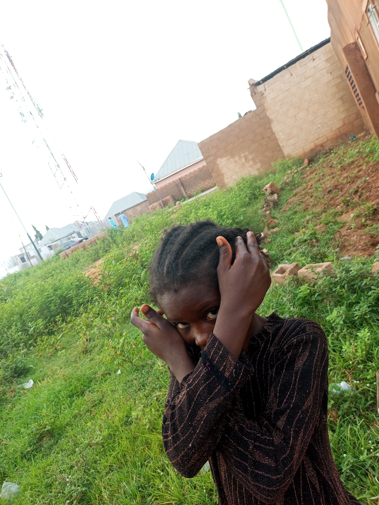 A young Black girl with dark braided hair is positioned in the foreground, looking directly at the camera with a shy expression, her hands held up to her face. She is wearing a long-sleeved, dark-colored top with a patterned design. Behind her, a slightly sloping landscape is covered in green grass and scattered with some small rocks and building materials. In the midground, there are several unfinished brick buildings and structures, typical of a developing area. Further in the background, more residential buildings with pitched roofs and a large telecommunications tower are visible under an overcast sky. The scene appears to be an outdoor location in the city of Tempure, Nigeria, characterized by its rural or semi-urban development.