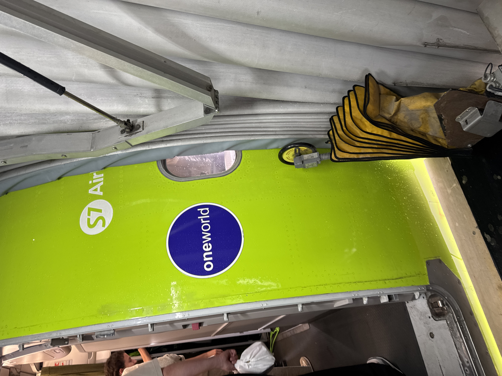 The image shows a close-up of an airplane door, painted bright green, with the S7 Airlines logo visible. A "oneworld" alliance logo is also present on the door. The door appears to be open, revealing the interior of the aircraft. To the right of the door, there is a gray, corrugated wall and a metal support structure with a gas strut attached. A yellow bellows-like object with black trim is resting against the wall. In the background, on the left side of the image, a person is visible, holding a white plastic bag and walking into the aircraft. Another person's arm and shoulder are also visible. The floor inside the aircraft appears to be dark gray. The overall scene suggests an airport environment, likely at Pulkovo Airport (LED) in Saint Petersburg, Russia, given the S7 Airlines branding.