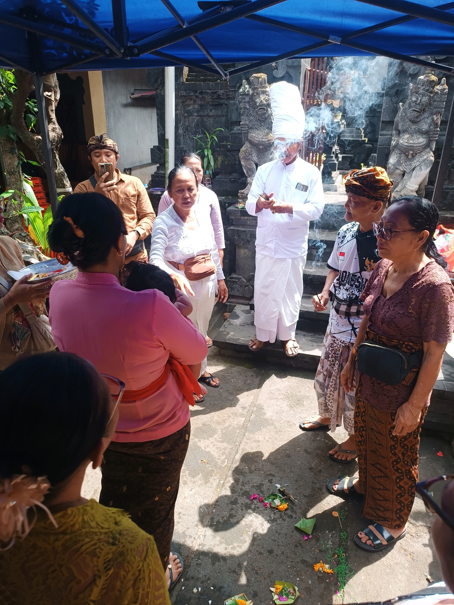 A group of people are gathered outdoors, possibly for a ceremony, under a blue tarp. In the foreground, a woman in a pink top and brown patterned sarong is holding a baby. To her left, another person in a yellow top is partially visible. In the middle ground, a man in a brown shirt holds a phone to his ear, while a woman in a white outfit stands beside him. Further back, a man in a white robe and tall white head covering is holding incense, with smoke rising around him. He stands between two stone statues. To his right, a man in a black and white shirt with Indonesian flag motif and a patterned sarong stands near a woman in a brown lace top and patterned sarong, who is wearing glasses. Offerings of flowers and greenery are scattered on the ground. The background suggests a traditional stone structure. The scene is set in Tabanan, Indonesia.