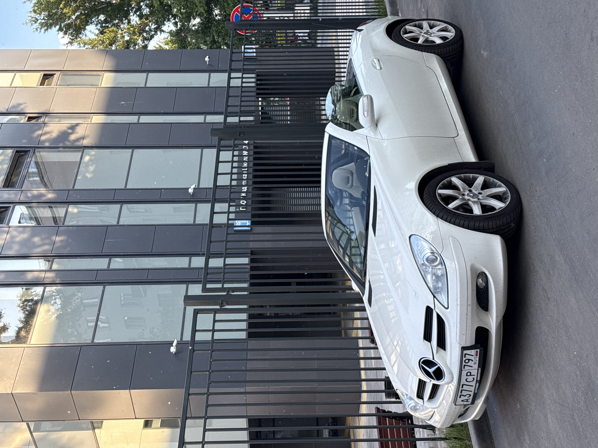A white Mercedes-Benz SLK-Class convertible is parked on an asphalt road next to a modern building in an urban setting. The car, a two-door model with alloy wheels, displays a Russian license plate "A377 CP 797 RUS" on its front bumper, with "797" indicating the Moscow region. Additional text "O2-PREMIUM | DOROFILOV" is visible on the license plate frame.
The building features a contemporary design with dark grey or black panels and large, reflective glass windows. Several small white security cameras are mounted on its facade. A black metal fence, composed of horizontal bars, runs parallel to the building and the car. Mounted on the fence is a sign with Cyrillic text that reads "ПОД'ЕЗД № 7 4" (Entrance No. 7 4) and a blue square sign. Behind the fence, further into the background, a round red and blue prohibitory road sign is partially visible.
The scene is captured during daytime, under bright sunlight, which casts shadows and creates strong reflections of trees and the clear sky on both the car's shiny surface and the building's windows. Green foliage from trees is visible at the top of the image. No people or specific activities are depicted; the car appears to be stationary.