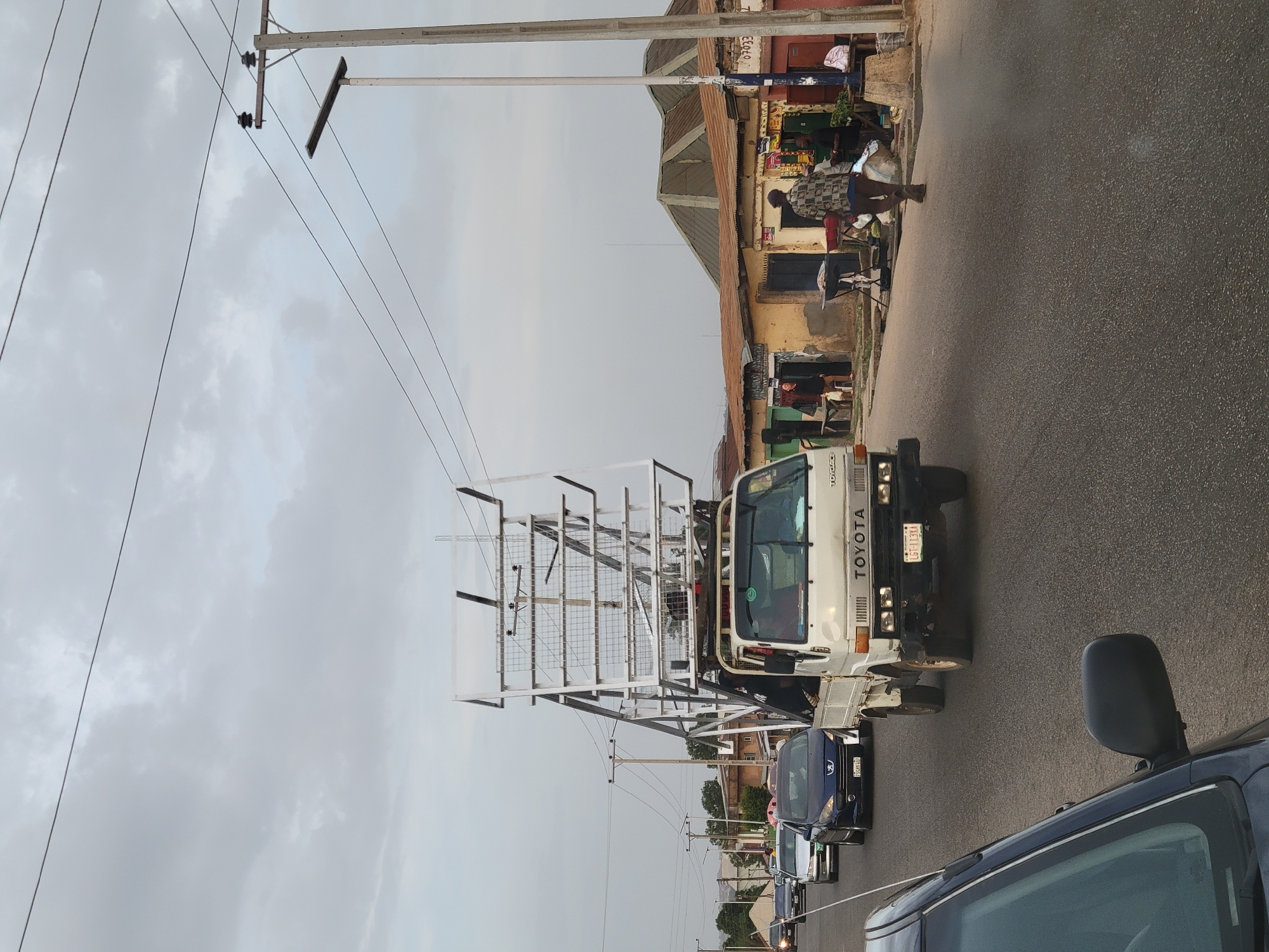 The image depicts a street in Jos, Nigeria, characterized by overcast skies and a road lined with buildings. A white Toyota truck with a large metal framework mounted on its bed is prominently featured, moving along the road. Several other vehicles, including dark-colored cars, are lined up behind the truck. Utility poles with power lines are visible along the roadside. A person is walking along the edge of the road near a building that appears to house small shops or businesses. The street is paved, and there is a sense of typical urban activity.