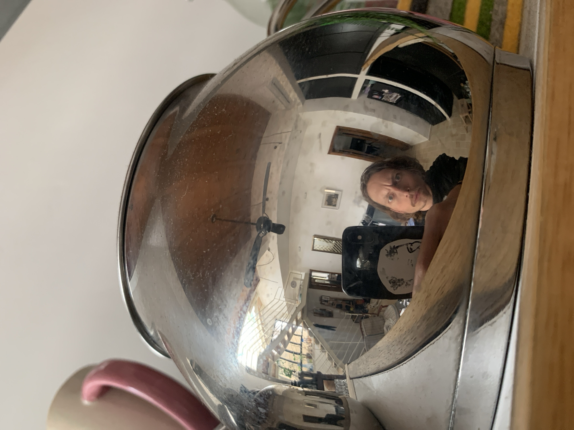 A distorted reflection is visible in a polished metal object, likely a pot or bowl. The reflection captures a room with a stairway leading to an upper level. Windows are visible, suggesting natural light. A person holding a phone is reflected in the metal object.
There are no indications of the location being Tel-Aviv or Israel in the reflection.