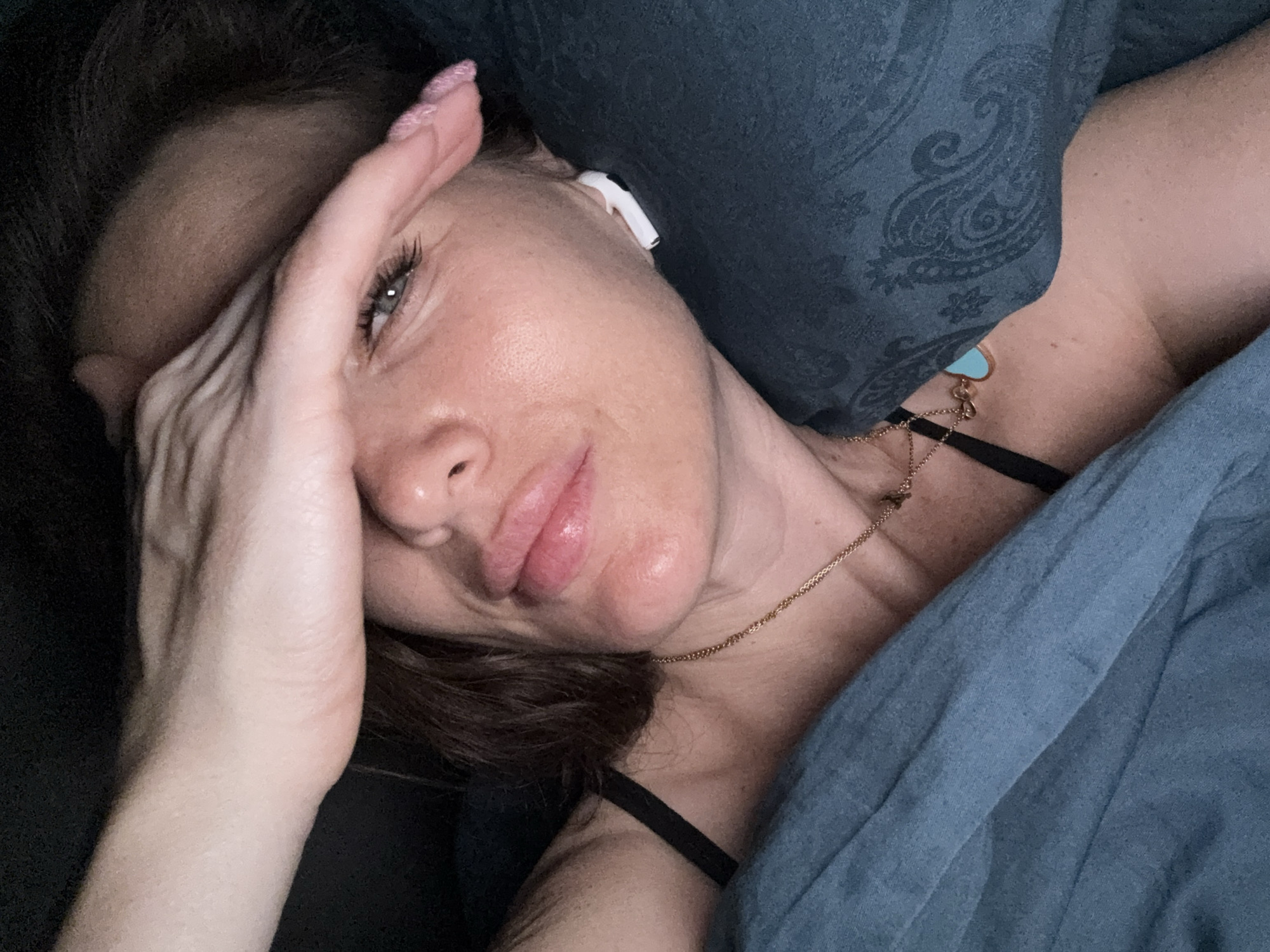 A woman lies in bed with her hand partially covering her face. She is wearing an earbud in her left ear and a gold necklace with a blue heart pendant. She is covered with a blue blanket and pillow. There is no indication of the location being Khimki, Russia.
