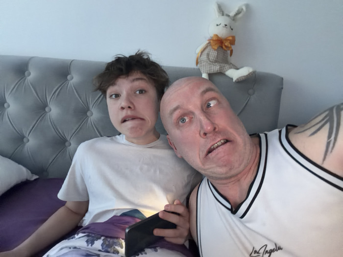 The image depicts an indoor scene with two individuals in what appears to be a bedroom in Chaikovsky, Russia. The precise location within Chaikovsky is not ascertainable from the visual information.
On the left, a younger male, with dark, messy hair, is seated, wearing a white short-sleeved t-shirt and patterned purple and white bottoms. He holds a black smartphone with an illuminated screen in his right hand. His facial expression is exaggerated, with pursed lips and wide eyes directed towards the camera.
On the right, an adult male with a bald head is positioned, wearing a white sleeveless tank top with "Los Angeles" printed on the front and black trim. He exhibits an exaggerated facial expression, with his mouth open, teeth visible, and eyes rolled upwards and to the right. A prominent black, possibly tribal-style, tattoo is visible on his left upper arm.
Behind the individuals is a grey, upholstered headboard featuring a tufted, diamond-patterned design with buttons. Perched atop the headboard, on the right side, is a white plush rabbit toy wearing a beige plaid vest and an orange bow tie. A light-colored wall is visible in the background. Below the younger male, a purple bedsheet and a portion of a white pillow are discernible. The overall scene suggests a posed, informal photograph.