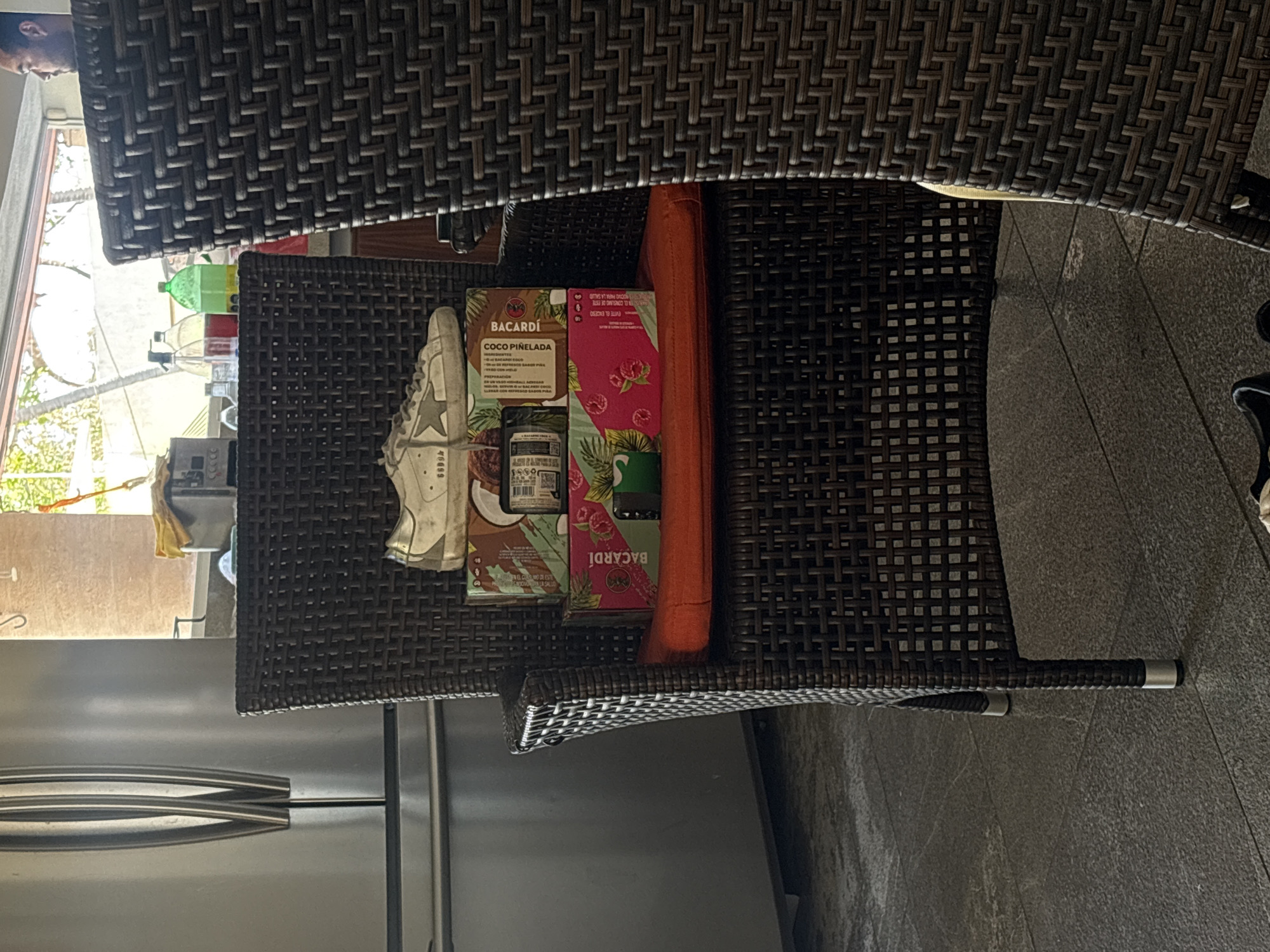 The image shows a close-up, angled shot of an outdoor setting, possibly in a residential or leisure area, that may be in Puerto Escondido, Mexico. The primary focus is on a dark-colored wicker chair. Positioned on the chair are a pair of white sneakers, a bottle of Bacardi Coco Piña Colada mix, and a box of Bacardi Raspberry Daiquiri mix. Adjacent to the chair is a stainless-steel refrigerator, suggesting an outdoor kitchen or dining space. The floor is tiled. A table with a similar wicker pattern partially obscures the view from above. Light filters in through the window, revealing a partial view of the street with trees and buildings in the background. An individual's face is partly visible in the upper left corner.