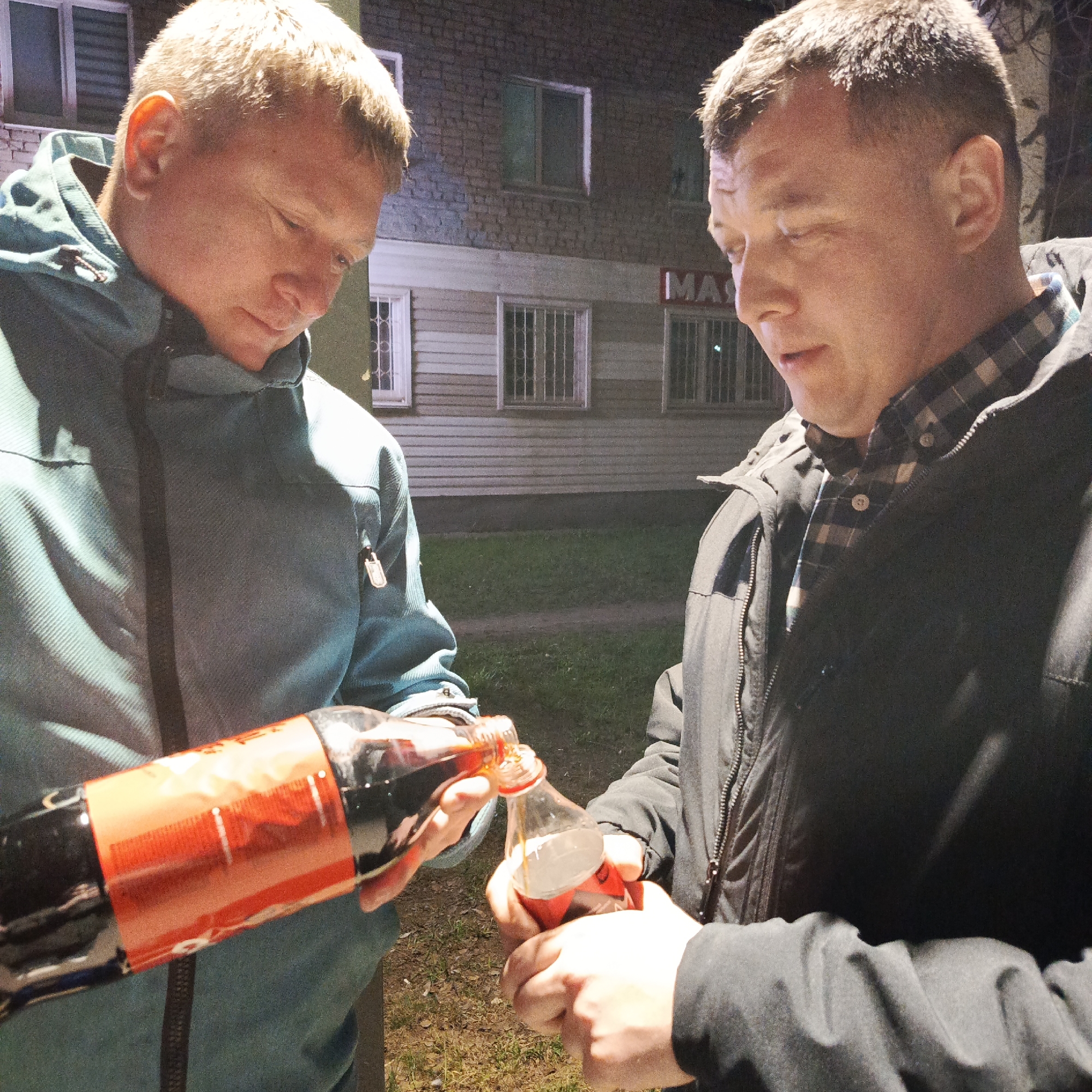 Two Caucasian males stand outside an unidentifiable commercial building in Chaikovsky, Russia. The building's facade is pale, with white-framed windows. One individual wears a blue hooded jacket, while the other is clad in a black jacket over a plaid shirt. The person in the blue jacket pours the contents of a large bottle, possibly a cola beverage, into a smaller bottle being held by the person in the black jacket. The scene is illuminated by artificial light, suggesting it is night.