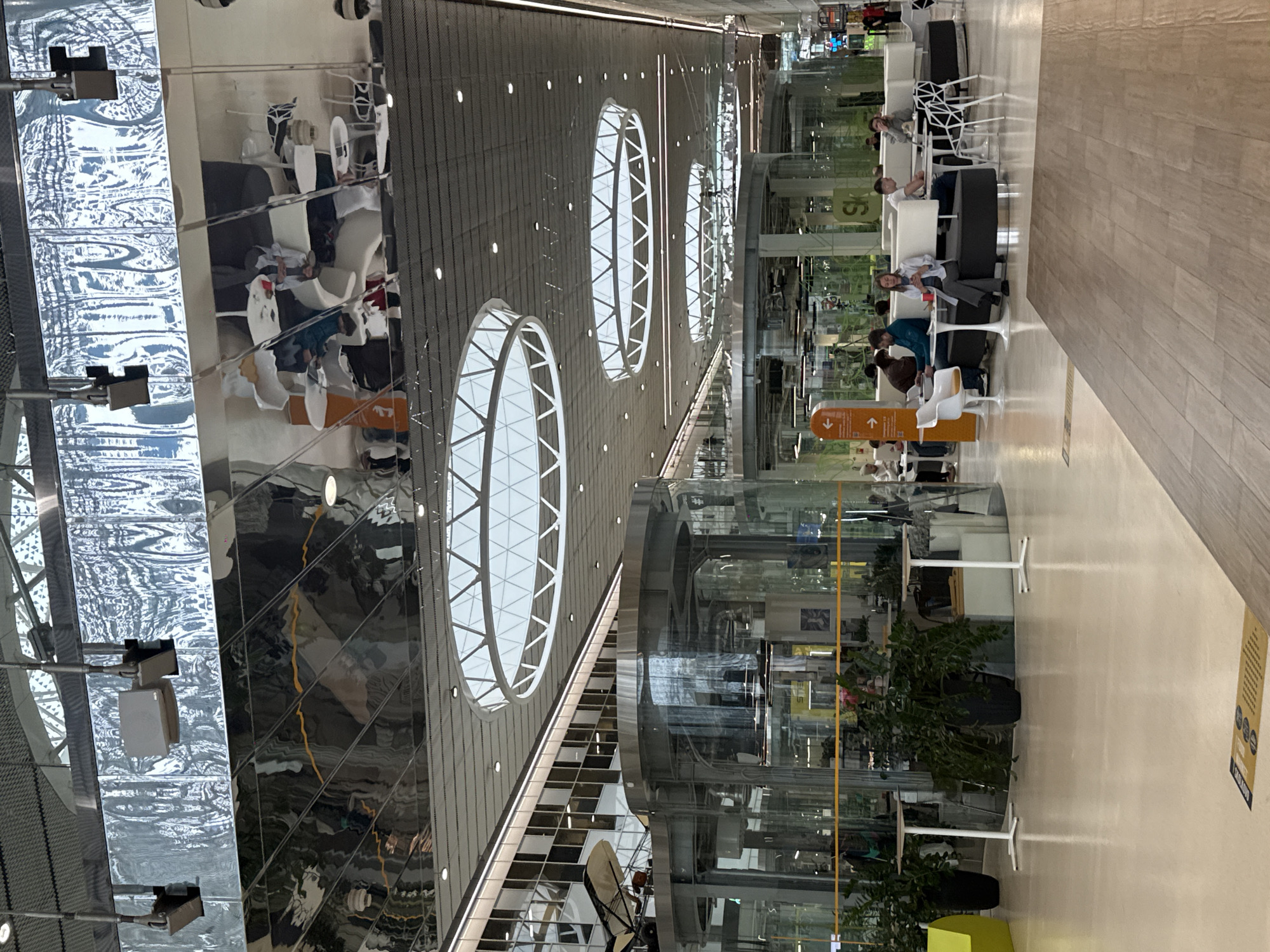 This image captures a spacious and modern indoor public area, likely an airport terminal or a large commercial complex, situated in Moscow, Russia. The scene is characterized by high ceilings featuring several large, circular skylights or decorative lighting fixtures, providing ample illumination and a sense of openness. The ceiling also integrates reflective panels and a textured, possibly metallic, surface with smaller recessed lights.
Multiple individuals are visible, seated in comfortable lounge-style seating areas on the right side of the frame. These areas are furnished with contemporary black and white sofas and chairs, accompanied by small white tables. The people appear to be casually dressed, engaged in resting, waiting, or quiet conversation.
On the far left, a highly reflective, corrugated or patterned wall surface dramatically mirrors and distorts the interior architecture and figures. In the center, large cylindrical glass structures, possibly enclosed escalators or decorative elements, rise through the space, further reflecting the surroundings. Potted green plants are strategically placed near the seating arrangements. The floor on the right exhibits a light-colored wooden pattern, complementing the sleek, modern aesthetic.
Visible text includes an orange directional sign featuring upward and downward arrows, and an icon resembling stairs or an escalator, with the word "TO." A yellow rectangular sign on the floor contains small, indistinct text and icons, potentially offering information or advertisements, one of which appears to be a "WiFi" symbol. Through the glass, parts of a green sign showing "SK" are discernible, possibly indicating a brand or service. The overall atmosphere suggests a well-designed, functional, and somewhat bustling public waiting or transit area.