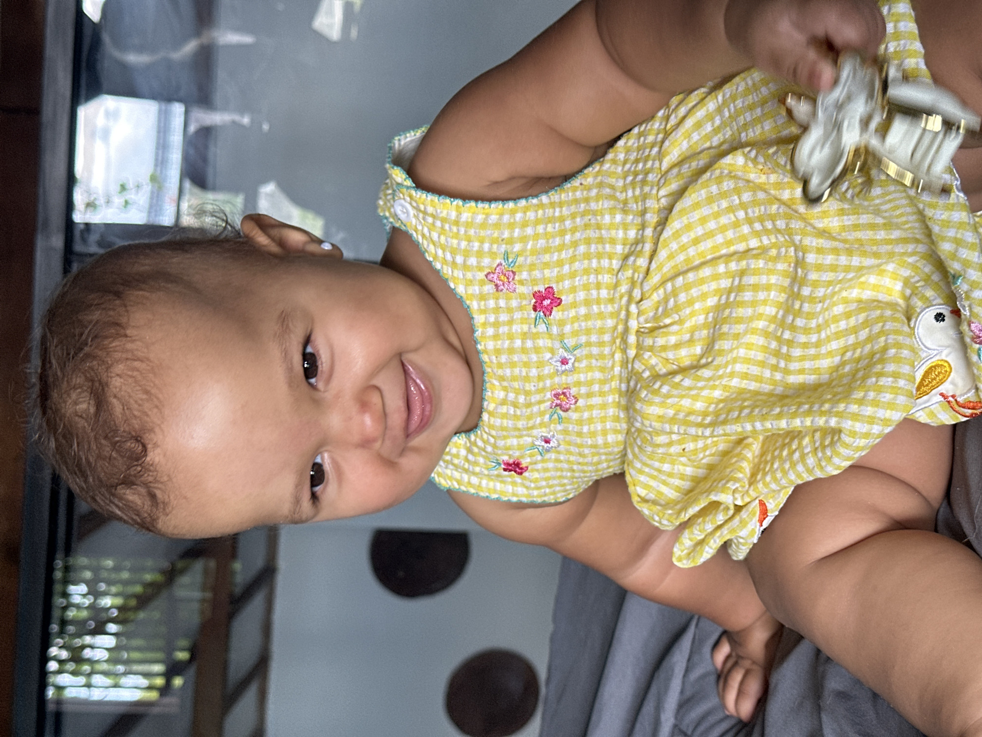 A young child, likely a toddler, is seated on a gray surface, possibly a bed or sofa. The child is wearing a yellow and white gingham dress with embroidered flowers on the front. The child is looking to the left with a wide smile and tongue sticking out. Their right hand is holding a small, light-colored object with gold accents, which is slightly blurred due to movement. The background appears to be an indoor setting with a blurred window or glass partition showing some greenery and reflections. The overall scene suggests a candid moment of the child at play. The location of North Kuta, Indonesia is not evident from the visual information in the image.