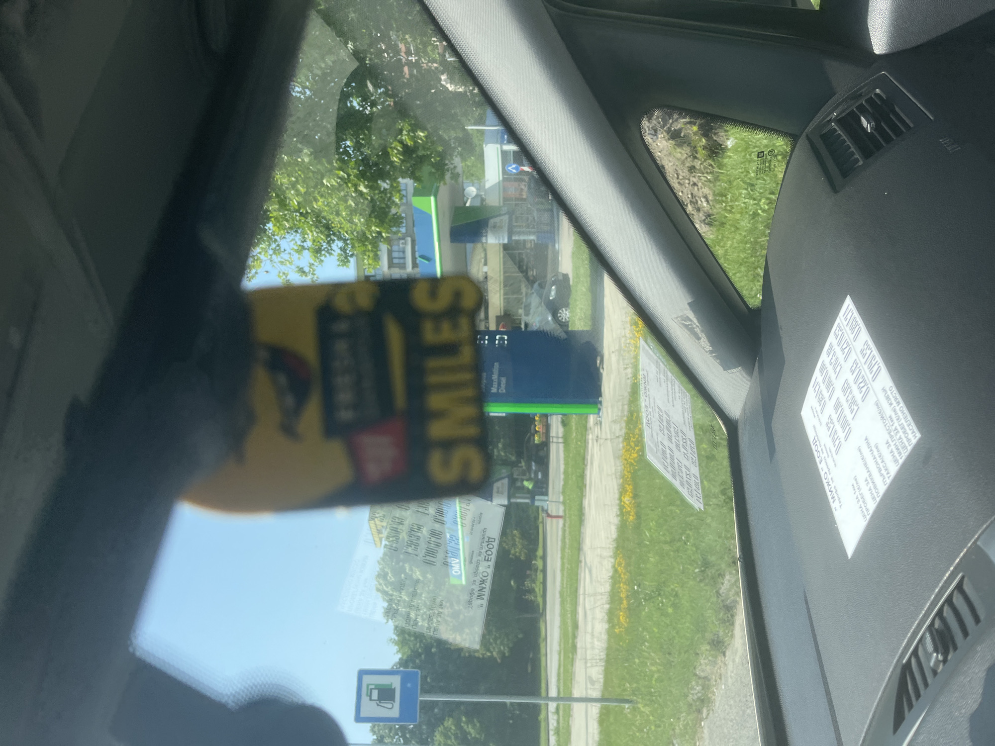 The image, taken from inside a car in Varna, Bulgaria, looks out through the windshield on a clear, sunny day. On the left side of the windshield, a yellow sticker partially obstructs the view, featuring the word "SMILES" and other partially visible text like "FRESH &..." and "MaxxMation Diesel".
Affixed to the dashboard and lower part of the windshield are several white papers or stickers, filled with Cyrillic text. These appear to be pricing or tariff information, likely for a taxi or delivery service. Visible terms include "МИЖО" ЕООД (a company name), "ЦЕНА ЗА 1км" (Price per 1km), "ПРОБЕГ (КМ)" (Mileage), "ПОВИКВАНЕ(Б)" (Call/pickup fee), "ЦЕНА ЗА ПЪРВОНАЧАЛНА ТАКСА" (Initial fare), and "НАСЕЛЕНО МЯСТО" (Populated place), along with various numerical values such as "1583.09" and "178318".
Outside the car, a street lined with green grass and trees is visible. A prominent blue sign with a white fuel pump icon indicates the presence of a petrol station. Behind it, a building with distinctive blue and green elements, likely part of the station or an associated shop, can be seen. Another sign board in the background confirms "MaxxMation Diesel". No individuals are explicitly visible, but the setting implies human activity at the petrol station. The overall scene suggests the vehicle is stationary, possibly at a fuel stop.