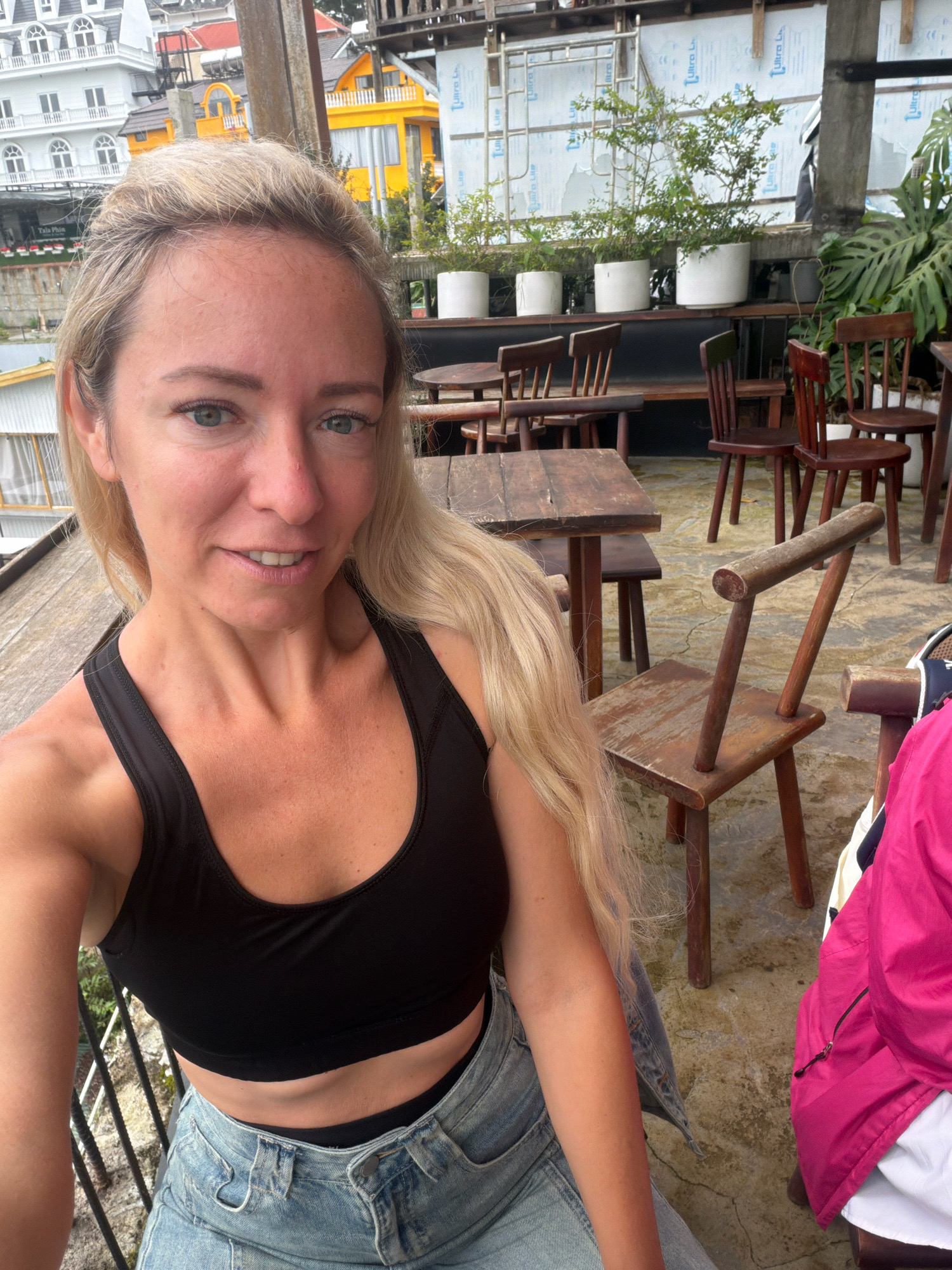 A blonde woman is taking a selfie from a seated position on a rooftop terrace in Da Lat, Vietnam. She is wearing a black sports bra and light blue denim jeans. Behind her, several wooden chairs and tables are arranged on a textured concrete floor. In the background, buildings with distinctive architecture, including white and yellow structures, are visible, along with potted plants. The overall impression is a casual, outdoor setting in an urban environment.