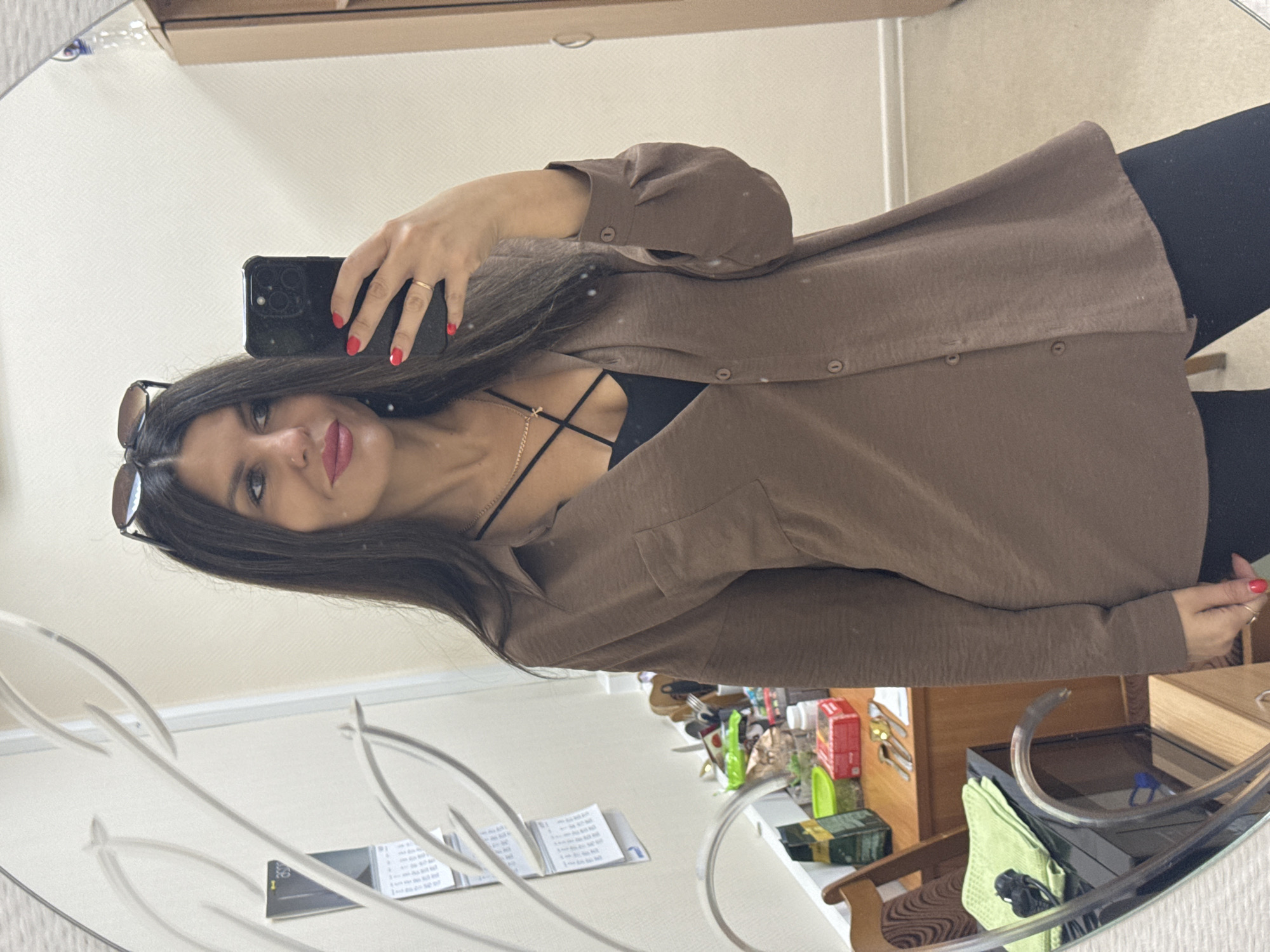 A woman with long dark hair and sunglasses on her head takes a selfie in a mirror. She is wearing a brown oversized button-up shirt with a black top underneath and a necklace. She has red nail polish on her fingers. She is standing in what appears to be a room with a desk and chair visible in the reflection of the mirror. The room has beige walls and a wooden floor. There are calendars and various items on the desk. The overall setting suggests an indoor personal space, possibly an office or a room in a home. There are no visual cues to suggest the specific location of Chaikovsky, Russia.