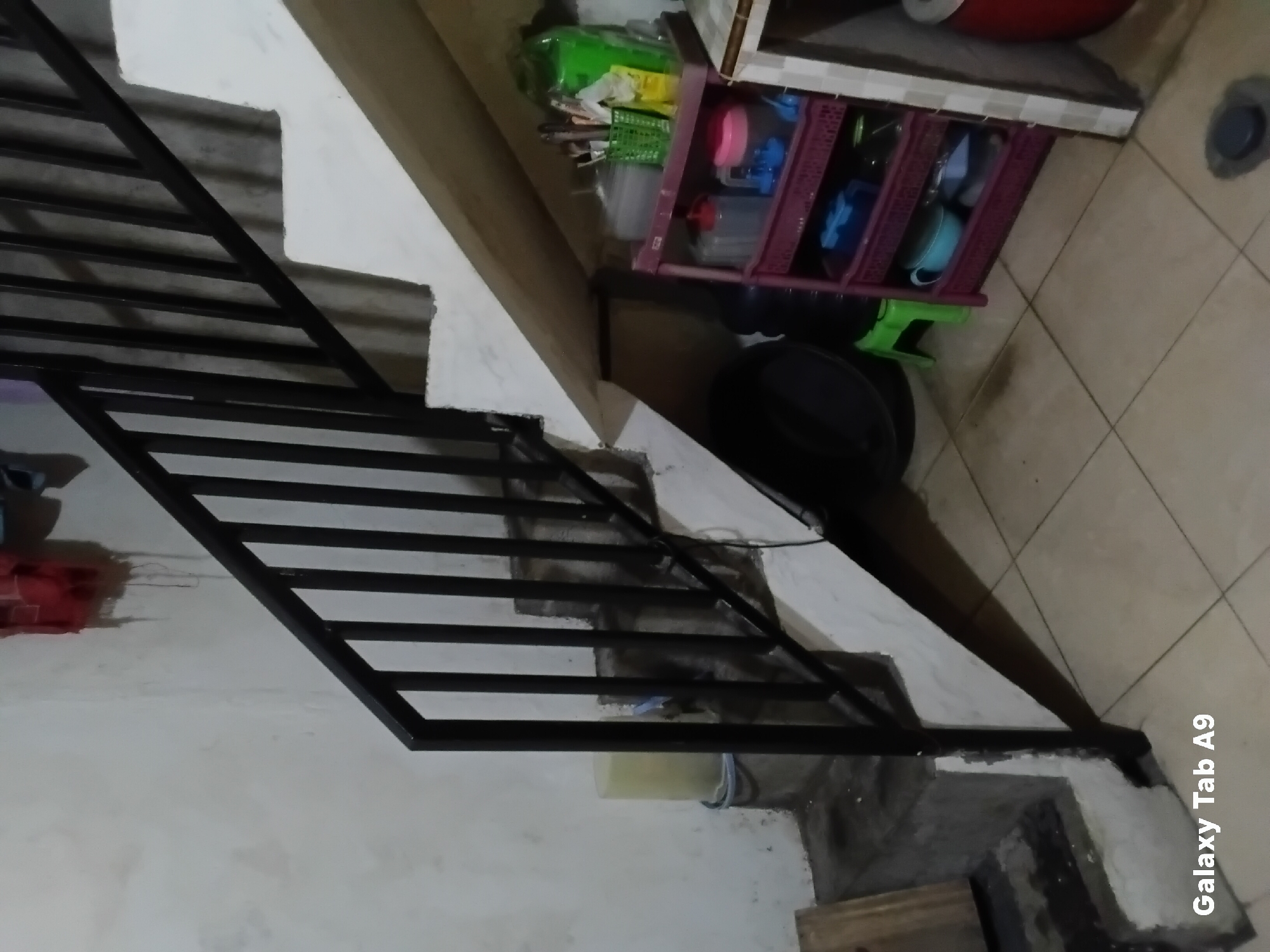 The image depicts a dimly lit indoor scene featuring a concrete staircase with a black metal railing. The stairs are partially visible and appear to be under construction or in a state of disrepair, with rough concrete surfaces. To the left of the stairs, a large pale yellow bucket sits on the floor. To the right of the stairs, a multi-tiered pink plastic shelf unit is filled with various household items, including containers, utensils, and what appears to be a green plastic basket holding more utensils. The floor is tiled with light-colored square tiles. A large black circular object, possibly a drum or container, is situated to the left of the shelf unit. In the background, a corrugated metal sheet is visible. The overall environment suggests a utilitarian or storage area. The bottom left corner of the image displays the text "Galaxy Tab A9".