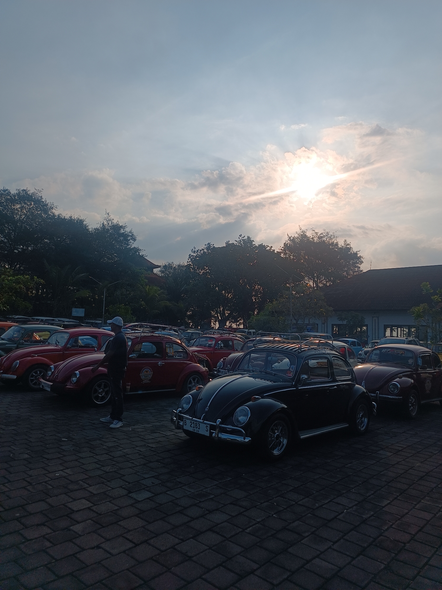 In the city of Kediri, Indonesia, a fleet of classic Volkswagen Beetles are parked on a cobblestone lot. The sun shines brightly through a cloudy sky, casting a warm glow on the scene. A man in a white cap stands between two red Beetles, appearing to inspect them. The cars are lined up, some with roof racks, and a few individuals can be seen in the background. Trees and a building with a dark roof are visible behind the vehicles, suggesting a park or event venue. The overall impression is of a car enthusiast gathering, possibly a local club meeting or exhibition.