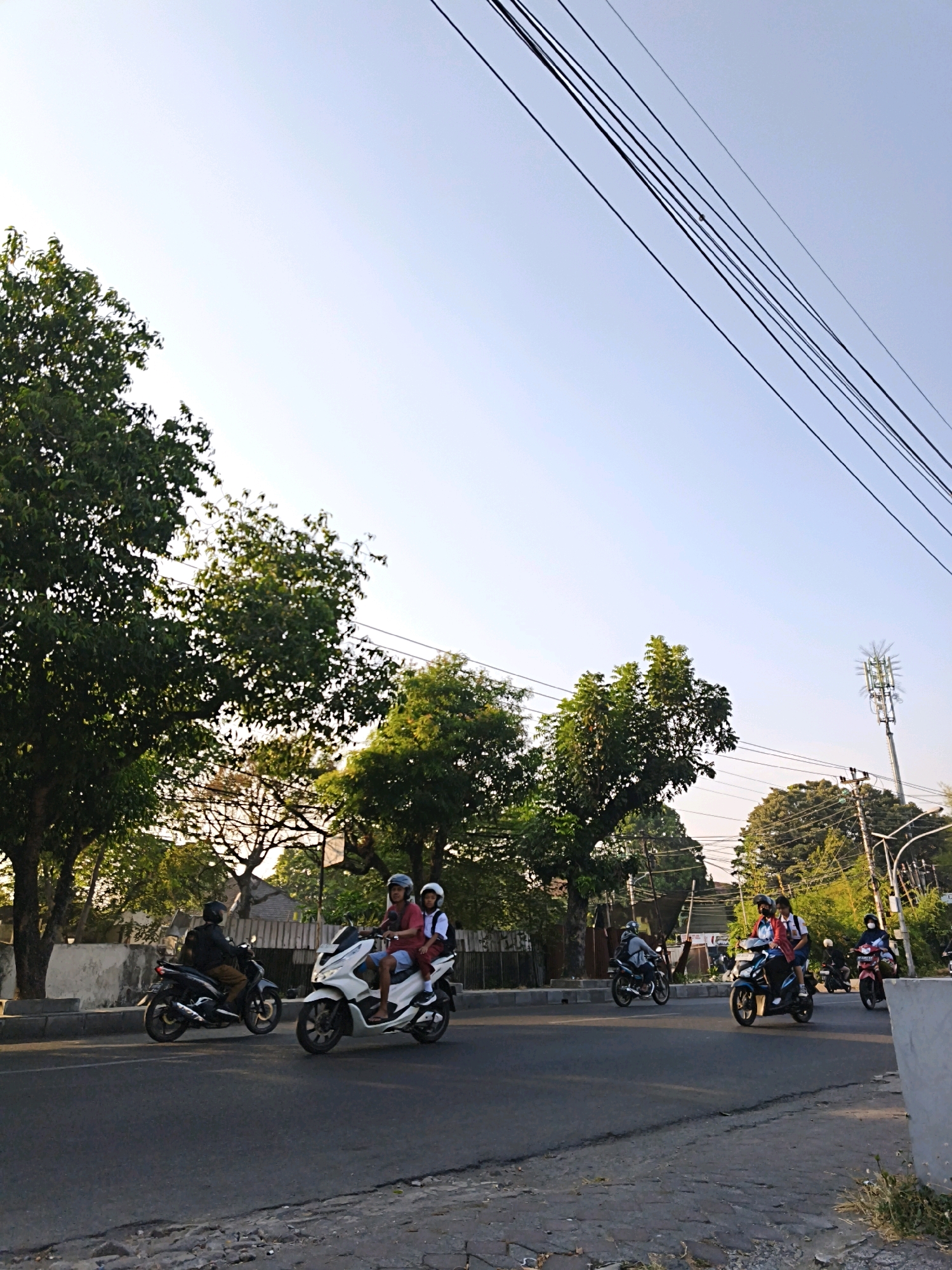 The image captures a street scene in Surakarta, Indonesia, under a clear, bright sky. A road with asphalt pavement runs horizontally across the frame, bordered by a raised curb and a sidewalk. Several trees with lush green foliage line the left side of the road, casting shadows on the pavement. In the background, more trees and buildings are visible.
The primary focus of the image is the traffic on the road. Multiple individuals are riding on motorcycles, moving in the same direction from left to right.
On the far left, a person on a black motorcycle is seen from the side. To their right, a white scooter is occupied by two individuals; the rider wears a red shirt and a helmet, and the passenger behind them wears a white shirt and a backpack, also with a helmet. Further down the road, another motorcycle with two occupants is visible, though details are less clear. Towards the right side of the frame, two more motorcycles are present, one dark-colored with two riders and another red and white scooter with one rider.
Overhead, a series of dark power lines are strung across the sky, and a tall telecommunications tower is visible in the distance on the right, surrounded by trees. The overall impression is a snapshot of daily life and transportation in a bustling urban environment in Surakarta.