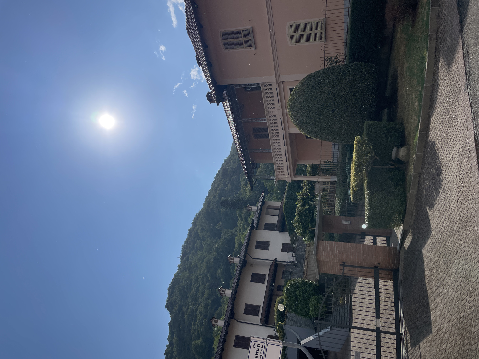 This image depicts a residential scene on a sunny day, likely in the city of Borgosesia, Italy, given the architectural style and lush, hilly terrain. In the foreground, a paved driveway with a herringbone pattern leads to a black metal gate. To the left of the gate, a sign reads "via Battisti." Adjacent to the gate is a brick pillar and a smaller pedestrian gate.
Two houses are visible. The house on the left is white with a tiled roof and brown shutters on its windows. It appears to be a row house or duplex with several units. The house on the right is larger and painted a pale pink color. It features a covered balcony with decorative railing, a tiled roof, and louvered shutters on its windows.
The landscaping includes neatly trimmed hedges, a large conical evergreen tree in front of the pink house, and a smaller, topiary-style hedge. Behind the houses, a steep, densely wooded hill rises under a clear blue sky, with the sun prominently visible and slightly overexposed in the upper right quadrant of the image. The presence of numerous small white specks in the air suggests dust, pollen, or perhaps insects.