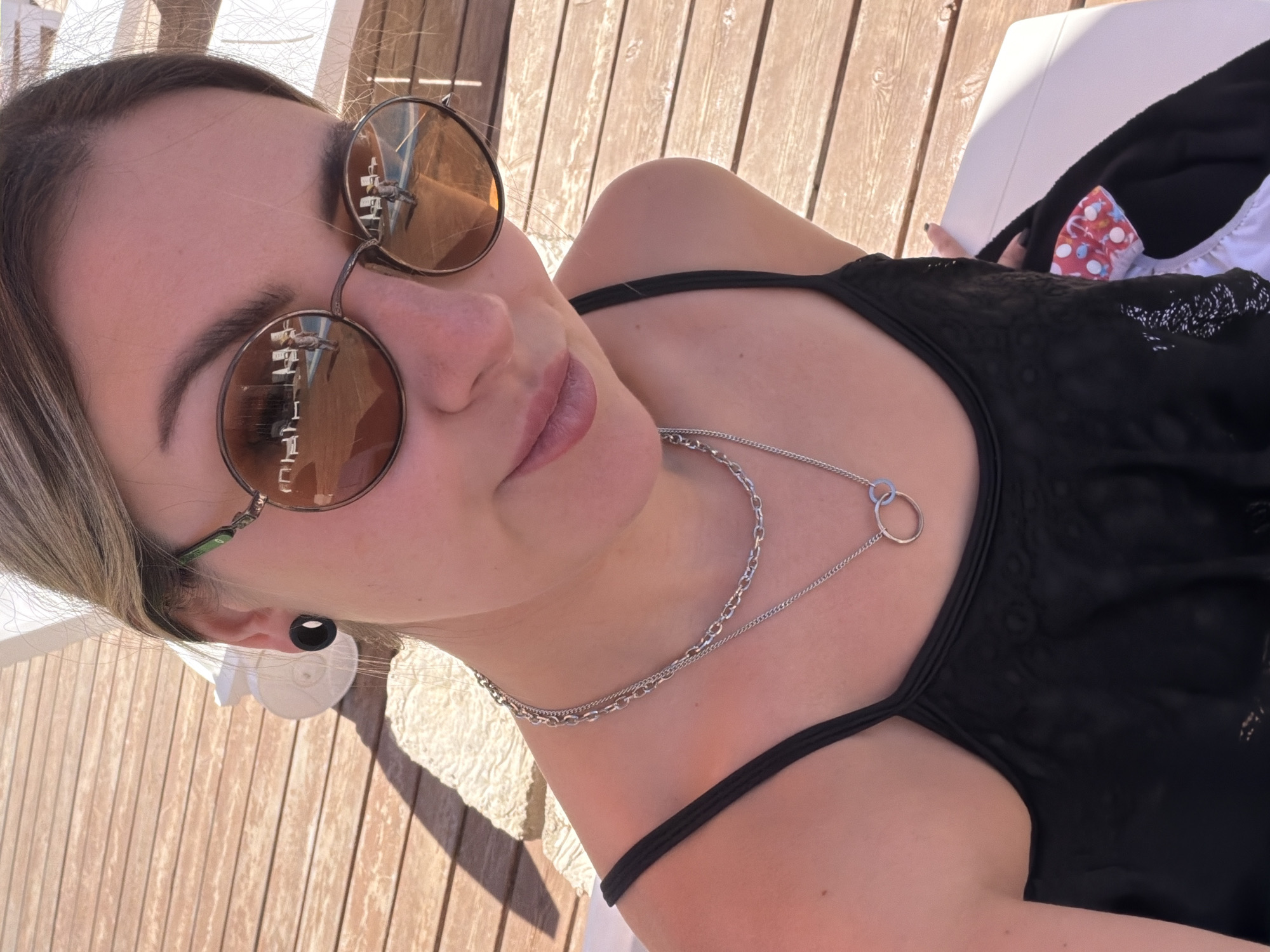 A young woman with blonde hair pulled back is captured in a selfie outdoors, sitting on a white lounge chair. She is wearing a black tank top and sunglasses with brown lenses. Her accessories include a black ear gauge and a silver necklace with two interlocking rings. The background consists of wooden decking and a glimpse of lounge chairs around a pool, suggesting a resort or recreational area. The sun casts shadows on the wooden surface.