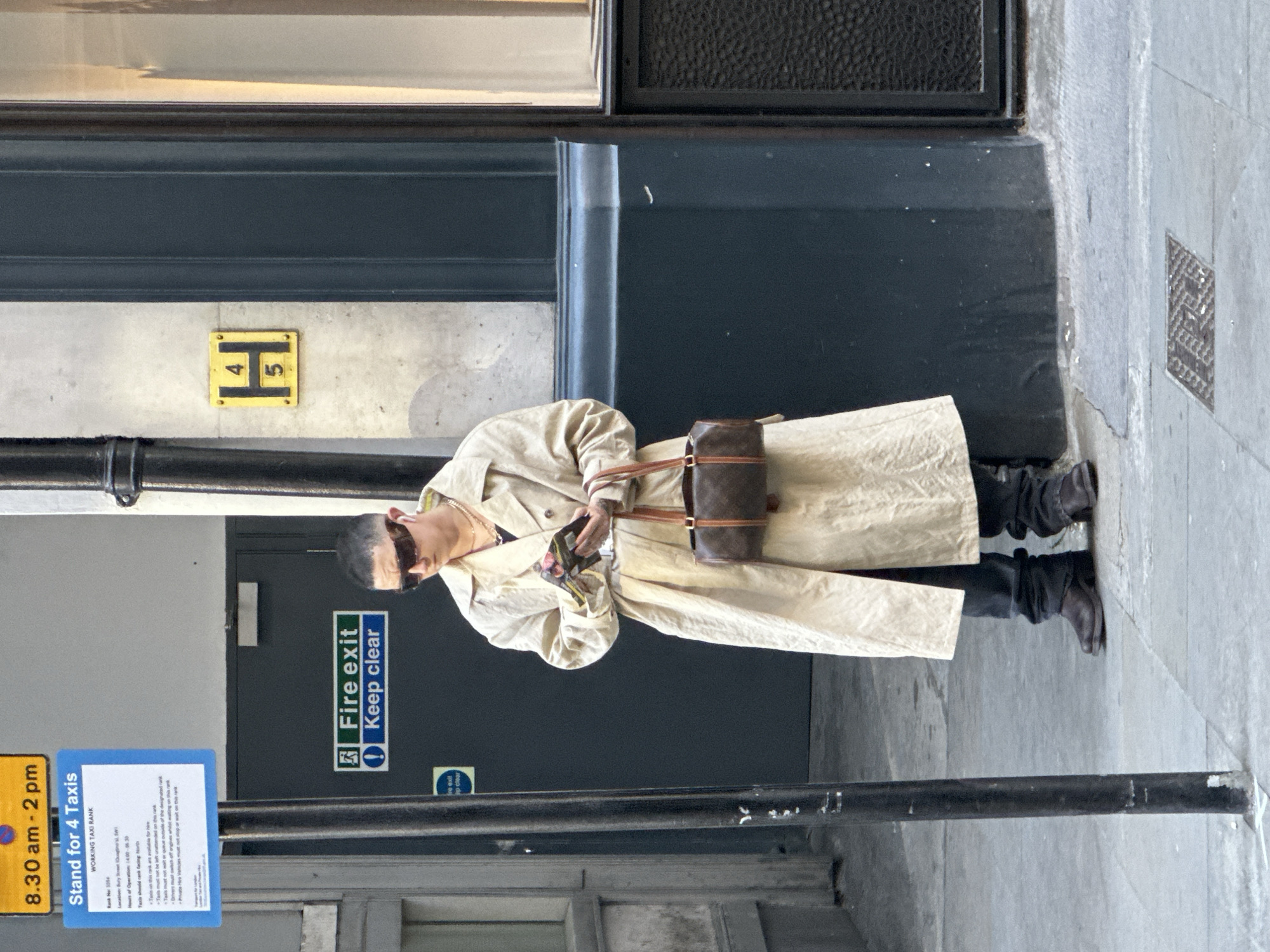 The image, currently oriented sideways, depicts an individual standing on an urban sidewalk, likely in London, United Kingdom. The person, appearing to be male, is dressed in a long, light beige or khaki trench coat, dark trousers, and dark boots. They wear sunglasses and have short, dark hair. In their hands, they hold a small, dark brown handbag with a light brown strap, and appear to be looking at something small held in their hands, possibly a phone or papers.
The background features a dark-colored building facade with a door. A green and white sign on the door clearly states "Fire exit" and "Keep clear". Above this, on the lighter-colored wall, is a small yellow square sign with a black 'H' and the numbers '4' and '5'. A black pole stands in the foreground, partially obscuring a black pipe running along the building's base. The ground is paved with grey concrete or stone slabs.
Attached to the pole is a blue and yellow sign for a taxi rank. Key visible text includes "8.30 am - 2 pm" and "Stand for 4 Taxis". Further details on the sign read "WORKING TAXI RANK", "Rank No: 1094", and "Location: Bury Street (Doughty St, SW1)", confirming the London setting. It also states "Hours of Operation: 14:00 - 06:30" and provides rules such as "Taxis should rank facing North" and restrictions on unattended taxis.
The scene appears to be set during the daytime under clear weather conditions. The individual's expression is obscured by sunglasses, but they seem to be calmly engaged with their activity.