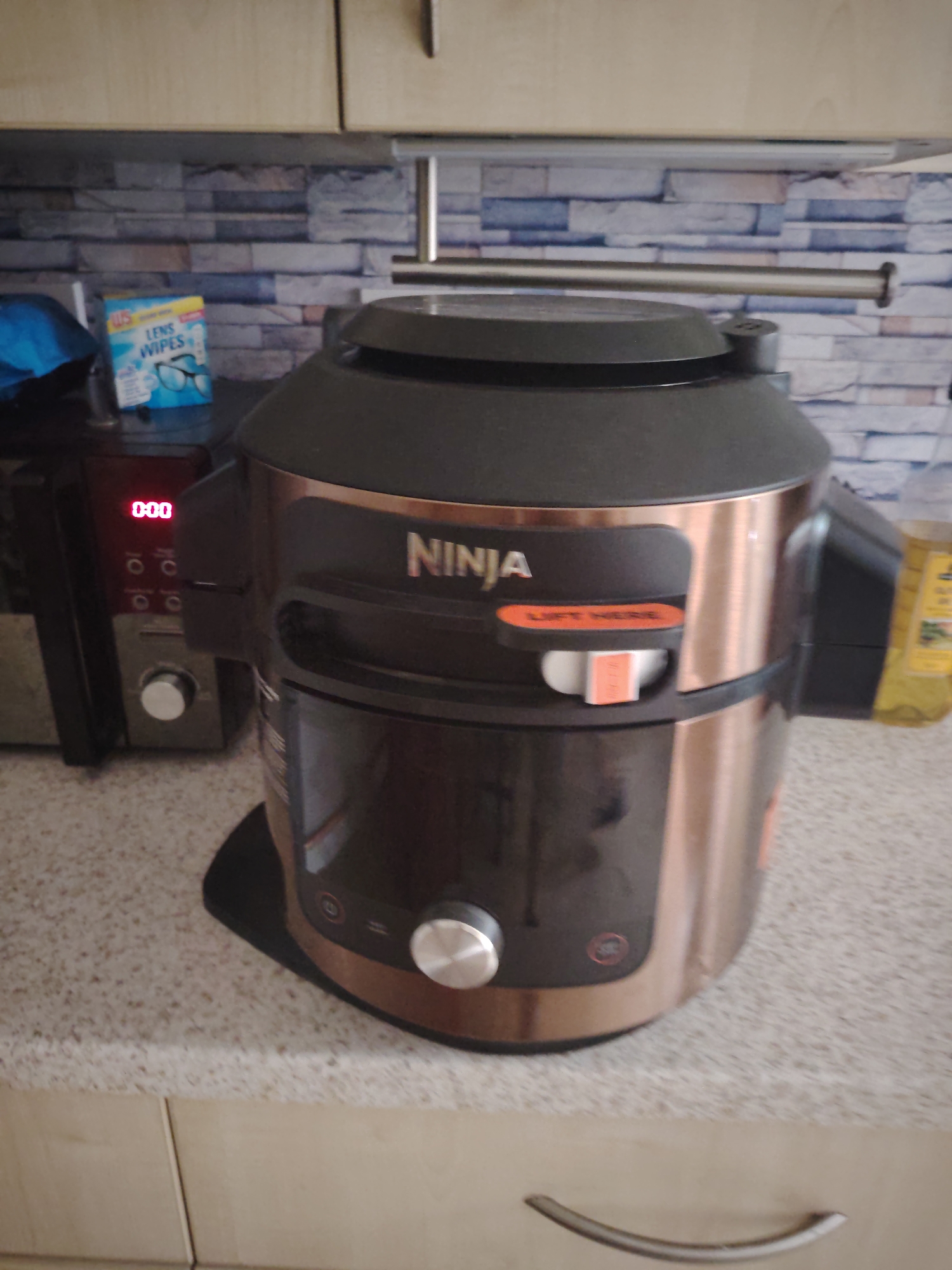 This image captures a kitchen countertop scene in Huntingdonshire, United Kingdom. In the foreground, a Ninja Foodi multi-cooker, colored in copper and black, is prominently positioned slightly right of center. The word "NINJA" is visible on its front, alongside an orange label that reads "LIFT HERE." A silver control dial is situated at the bottom front of the appliance. To the left of the Ninja appliance, a dark-colored kitchen appliance displays "0:00" on a red digital clock, with several control buttons and a silver knob below the display. Behind this appliance, a blue object is partially visible, and a box of "US LENS WIPES" featuring eyeglasses is visible. The background features a tiled backsplash with a textured, stacked stone-like pattern in shades of grey, blue, and brown. Above the backsplash, light-colored wooden kitchen cabinets are present, with a silver horizontal bar, possibly a towel rail, mounted on the wall. A yellow bottle, possibly cooking oil, is partially visible on the far right, behind the Ninja appliance. The countertop surface is a speckled, light-colored material. Below the countertop, light-colored wooden cabinet doors with a silver curved handle are visible. No individuals are present in the image.
