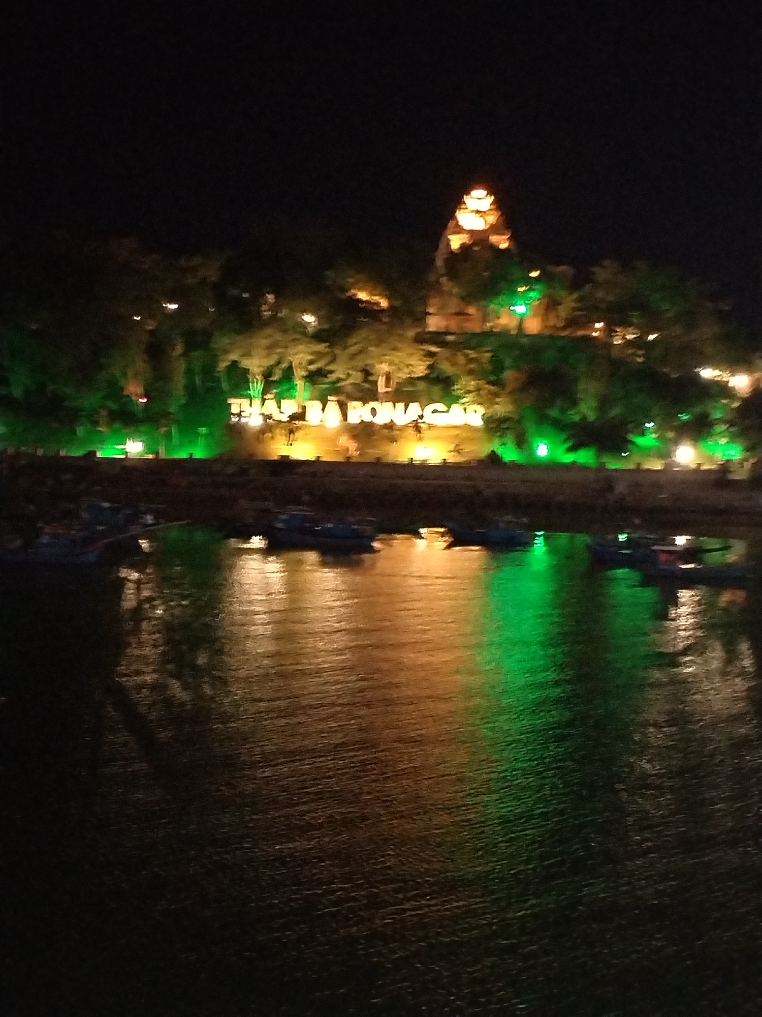 The image depicts the illuminated skyline of Tháp Bà Ponagar temple complex in Nha Trang, Vietnam, at night, as viewed from across a body of water. The temple's towers are lit in golden hues, while the surrounding vegetation and signage display green illumination. Several small boats are moored near the shore. The water reflects the lights, creating streaks of green and gold on the dark surface.