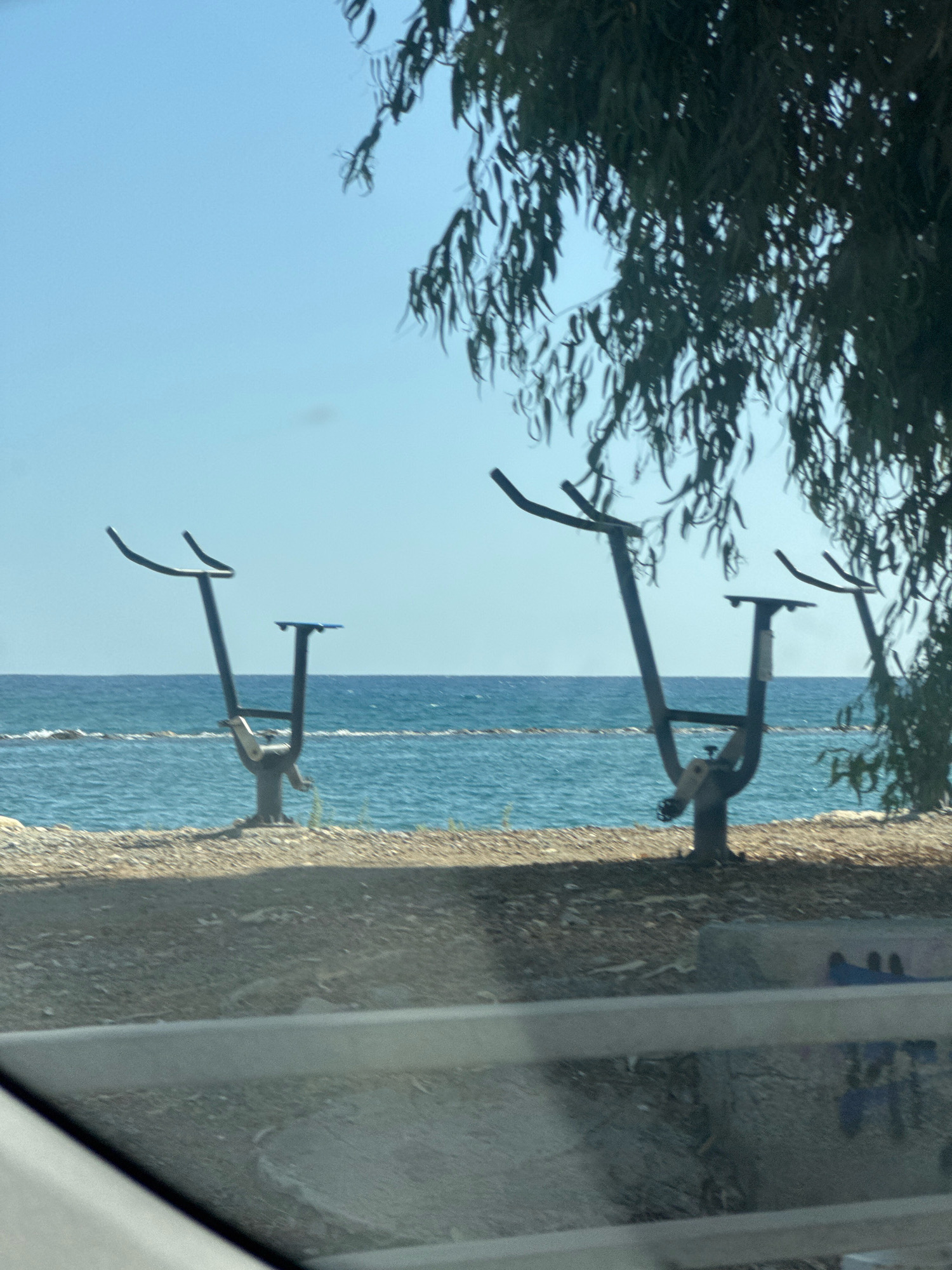 The image shows a scenic outdoor view with several pieces of exercise equipment situated on a beach next to the sea. The exercise machines appear to be elliptical trainers designed for outdoor use, with a dark metal frame and a blue seat. They are positioned along the shoreline, suggesting a public fitness area. The sea is visible in the background, with calm blue waters and a clear sky above. A large tree with long, drooping leaves casts a shadow over the scene, adding a natural element. The foreground shows a gravelly ground and a hint of a car's interior, indicating the photo was taken from a vehicle. The location is identified as Germasogeia Municipality, Cyprus, which is known for its coastal areas and recreational facilities.